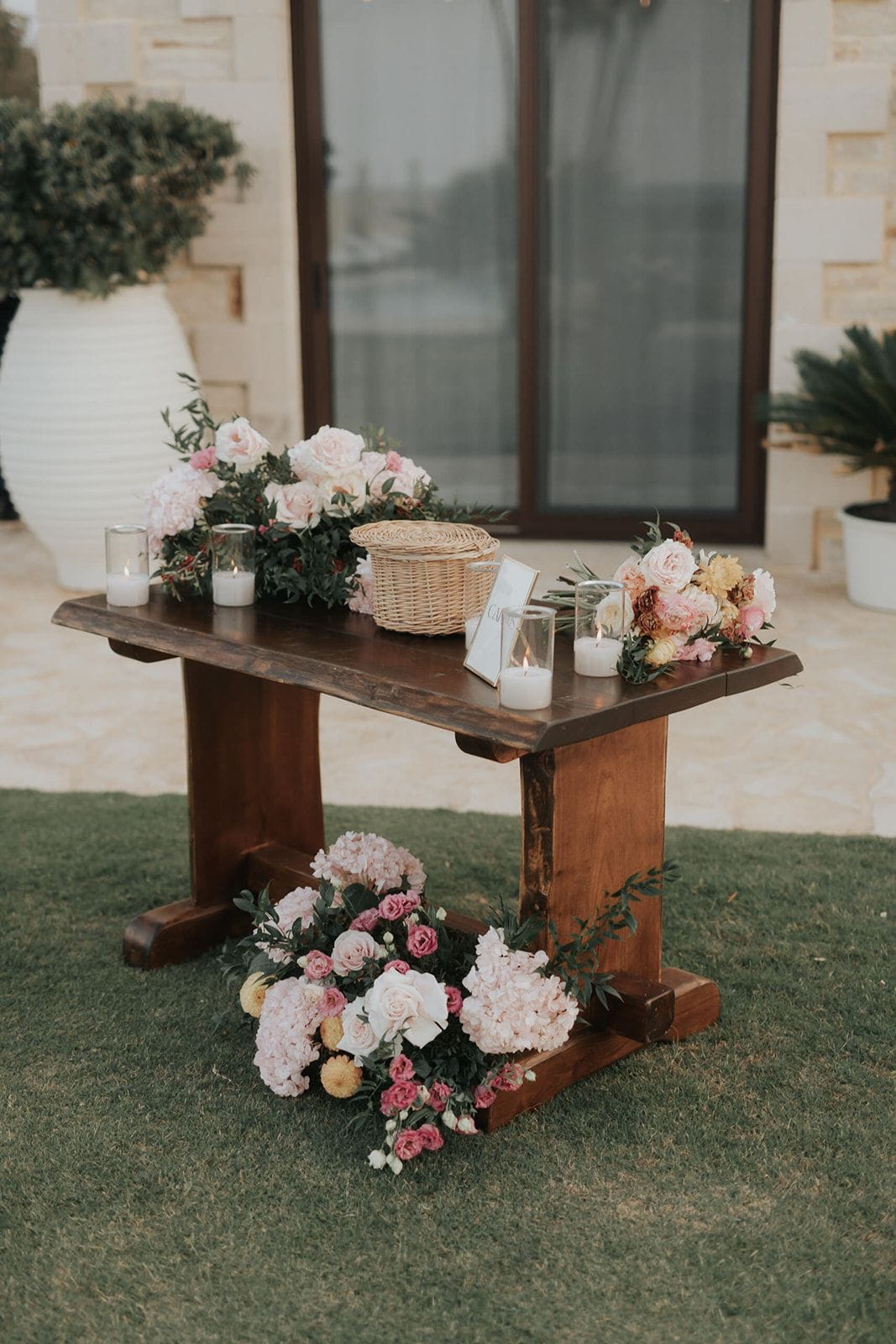 A rustic dark wooden table on a stone terrace holds a lush wedding arrangement of blush garden roses, pale pink hydrangea, pink spray roses and trailing olive and eucalyptus foliage. A woven wicker basket, glass hurricane candles and a small framed card sit among the flowers, and a second low cluster of pink and white blooms rests on the grass at the table's base. The soft, natural light gives a warm, romantic feel, set at Mitchell & Antoinnette's wedding in Greece.