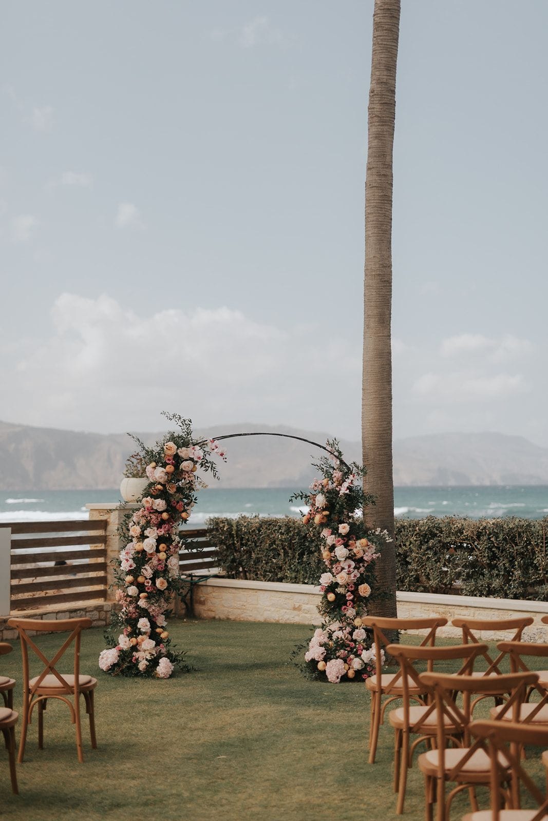 A wedding ceremony arch on a lawn overlooking the sea in Greece, where a curved black metal frame is wrapped in blush and peach garden roses, pale hydrangeas, pink blooms and round golden fruit, softened with trailing green foliage. Rows of wooden cross-back chairs with cushioned seats face the arch, and a tall palm trunk and a low hedge frame the view of turquoise waves and hazy mountains under a soft grey sky.