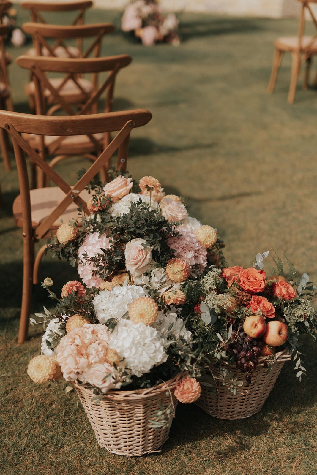 Two large wicker baskets overflow with wedding flowers set on grass beside a wooden cross-back chair: blush and white hydrangeas, pale pink roses, and round golden dahlias, woven through with dusty green foliage. The basket on the right holds coral-orange roses, apples, dark grapes and sprigs of eucalyptus, giving a warm autumn feel. These aisle arrangements, photographed outdoors at a wedding in Greece, are soft in colour and gently out of focus in the golden afternoon light.