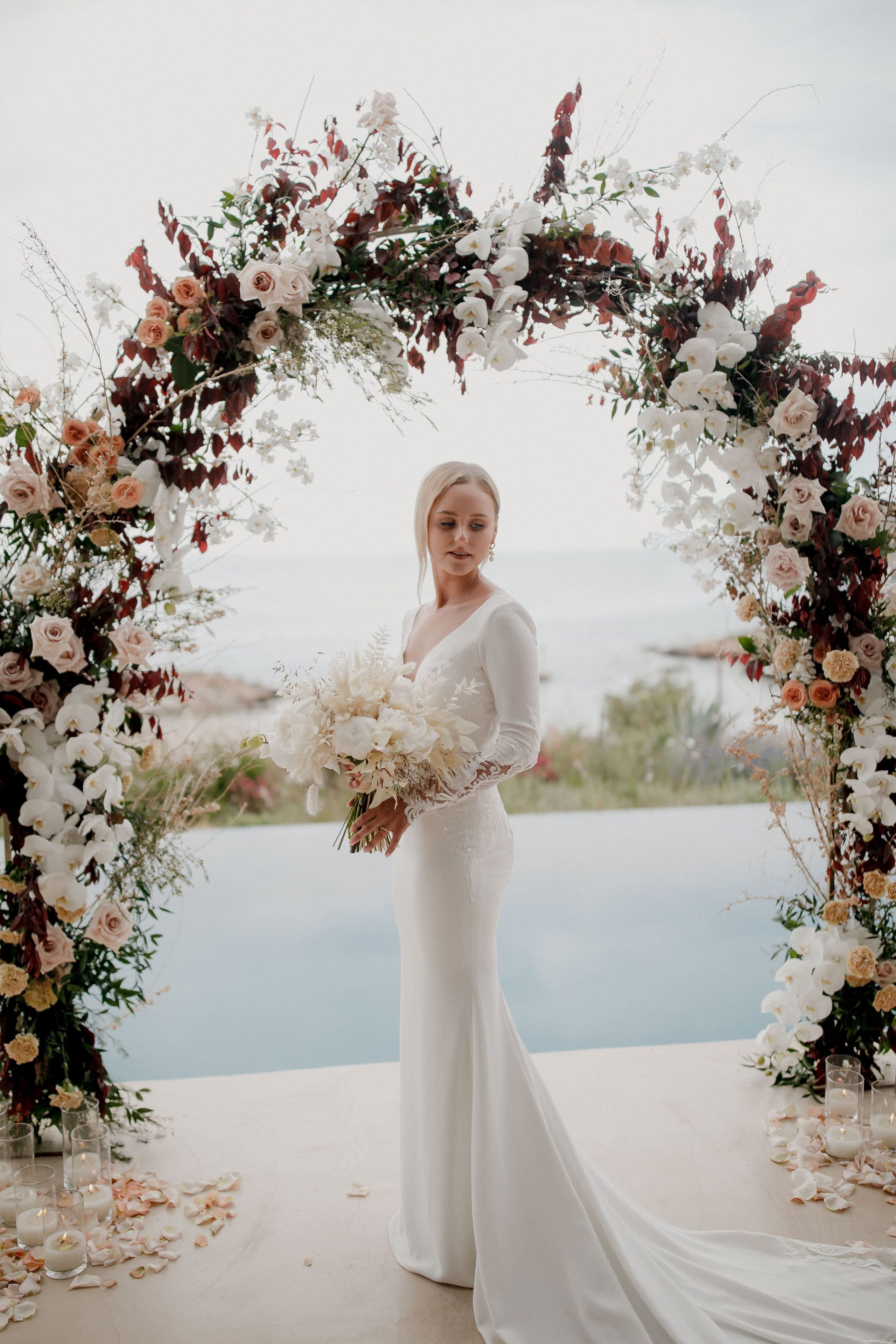 A bride in a long-sleeved, fitted satin gown with a sweeping train stands beneath a lush curved arch of blush garden roses, white orchids and burgundy foliage at Nuez Villas in Crete, Greece. She holds a cream bouquet of white blooms and looks down at it, with an infinity pool and the blue sea behind her, and petals and candles scattered along the floor. This is a wedding flower installation with a soft, romantic mood.