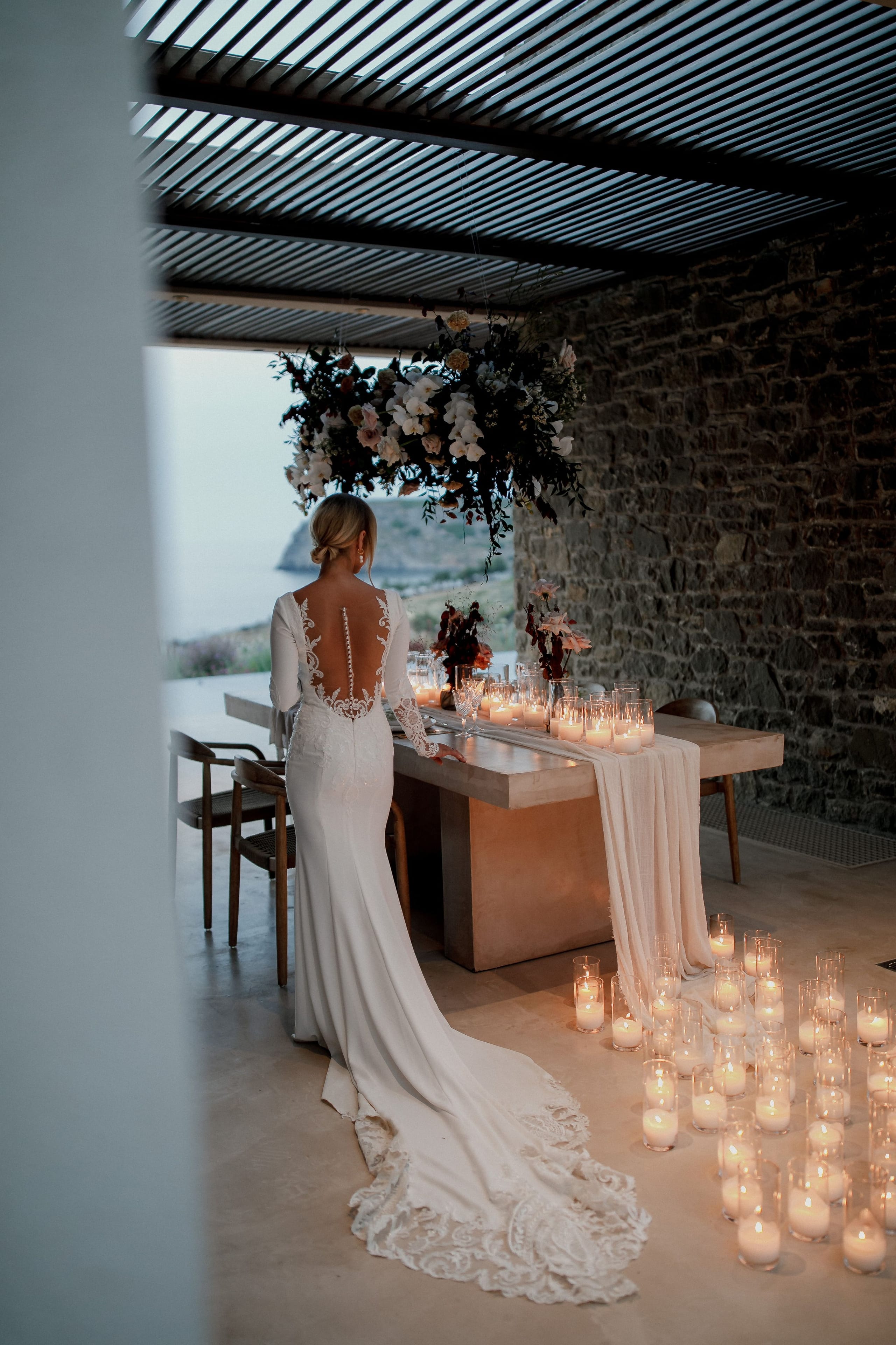 A bride in a fitted white gown with a sheer lace back and a long lace-edged train stands at a concrete dining table, facing a hanging installation of white orchids, pale pink blooms and dark foliage above it. Glass votive candles line the table and spill down the floor in a glowing trail, with a soft pink runner draped across the table. Through the open side of the stone-walled terrace at Nuez Villas in Crete, Greece, the sea and a rocky headland fade into dusk, creating a quiet, romantic wedding table setting.