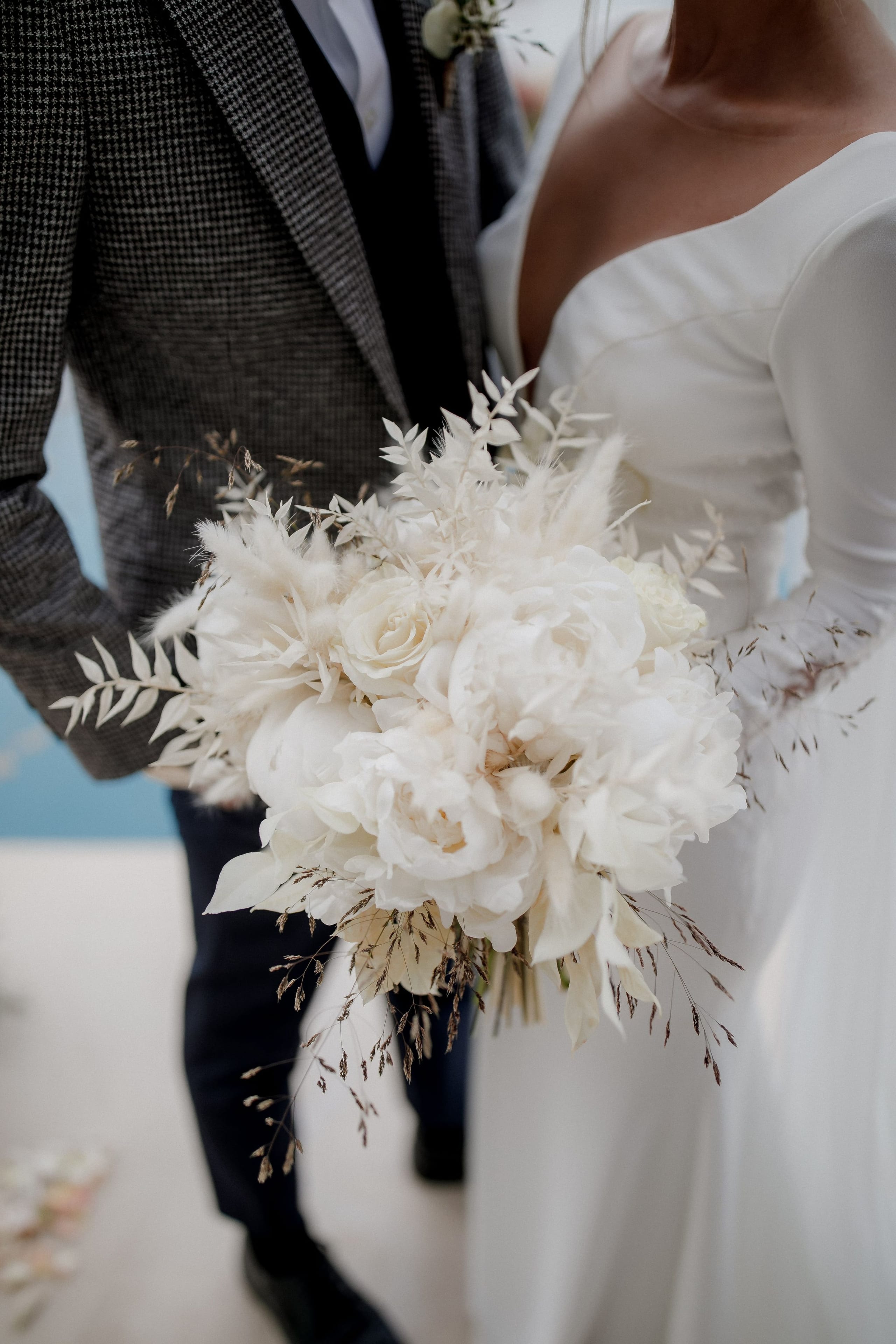 A close view of a bride's bouquet of creamy white peonies and garden roses, with wispy dried grasses, pampas-style plumes and feathery white foliage trailing from it. The bride wears a long-sleeved white satin gown with a sweetheart neckline, and beside her a groom in a grey houndstooth jacket holds her arm. This is a wedding bouquet at Nuez Villas in Crete, Greece, shot in soft natural light against a pale blue backdrop.