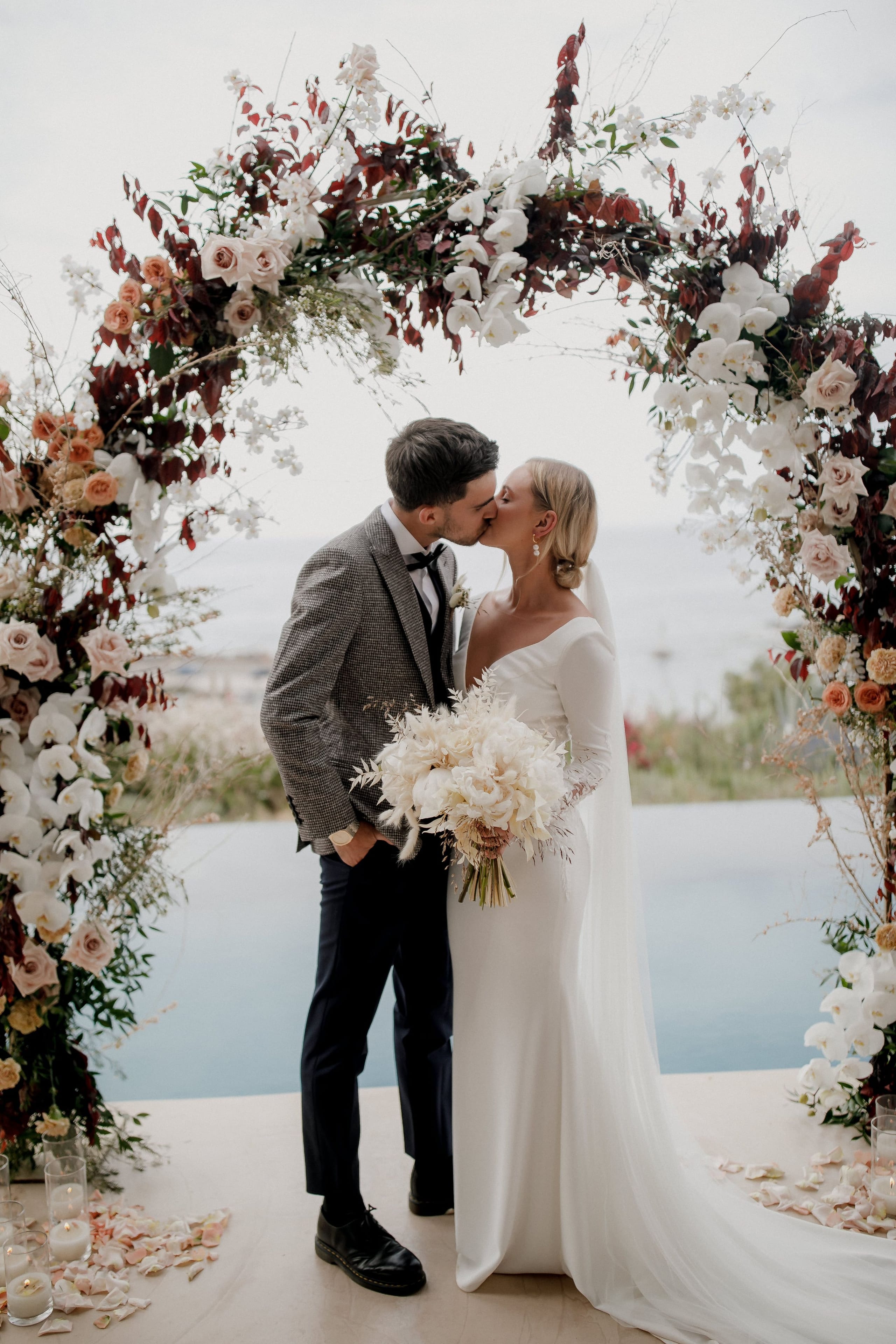 A bride and groom share a kiss beneath a tall, arching floral installation of blush roses, white orchids and deep burgundy foliage, with peach-toned blooms woven through, at the Nuez Villas wedding in Crete, Greece. The bride wears a fitted long-sleeved white gown with a flowing veil and holds a cream bouquet of full white and ivory blooms, while the groom wears a grey houndstooth jacket with a black bow tie. Behind them the sea glows pale blue, and rose petals and candles line the stone floor.