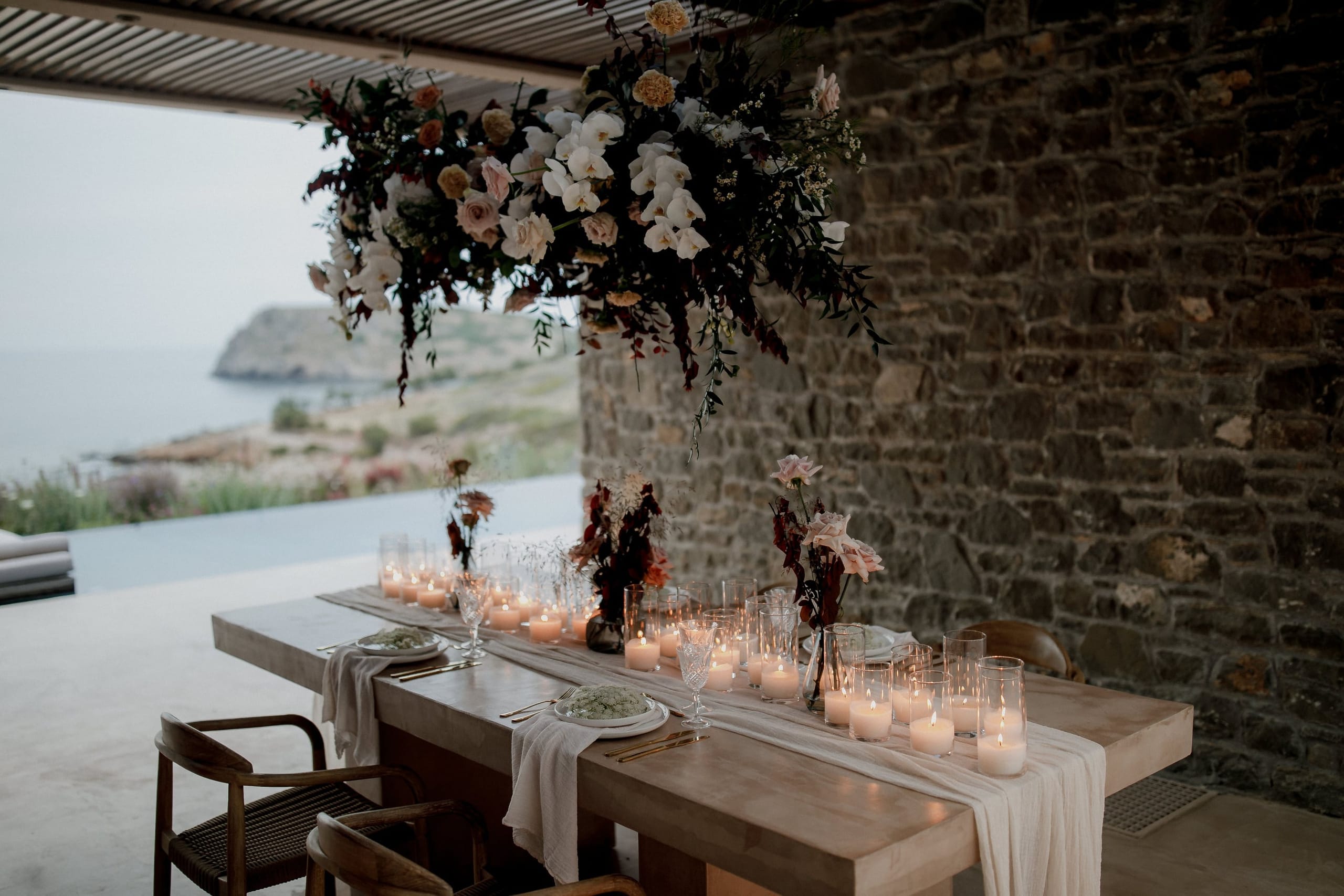 A long concrete dining table runs beneath a suspended cloud of wedding flowers, with cream and blush roses, white orchids and soft peach blooms trailing dark foliage above it. The table is dressed with a loose linen runner, rows of pillar candles in glass hurricanes and low vases of dusky blooms, set against a rough stone wall and an open view of the sea and rocky coastline at Nuez Villas in Crete, Greece.