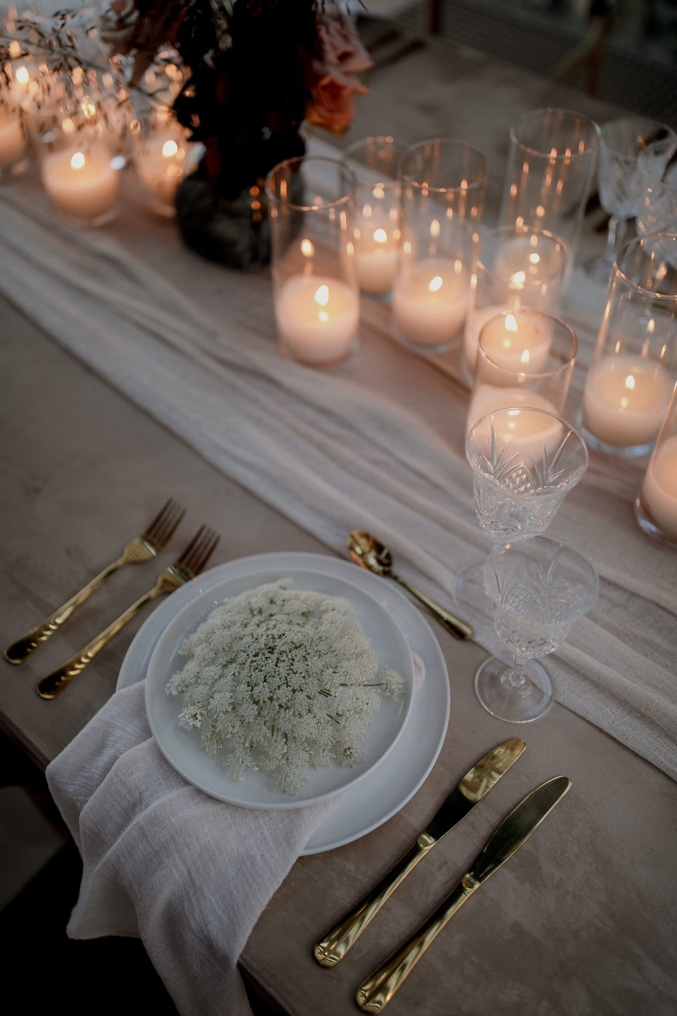 A wedding place setting at Nuez Villas on Crete, Greece, with a white plate holding a loose, lacy cluster of white cow parsley-style blooms on a pale linen napkin. Gold cutlery sits on either side, and a long gauzy runner stretches down the table, flanked by a row of glowing pillar candles in clear glass holders. Crystal wine glasses and a dark, moody arrangement of deep burgundy blooms in the background create a soft, intimate evening atmosphere.