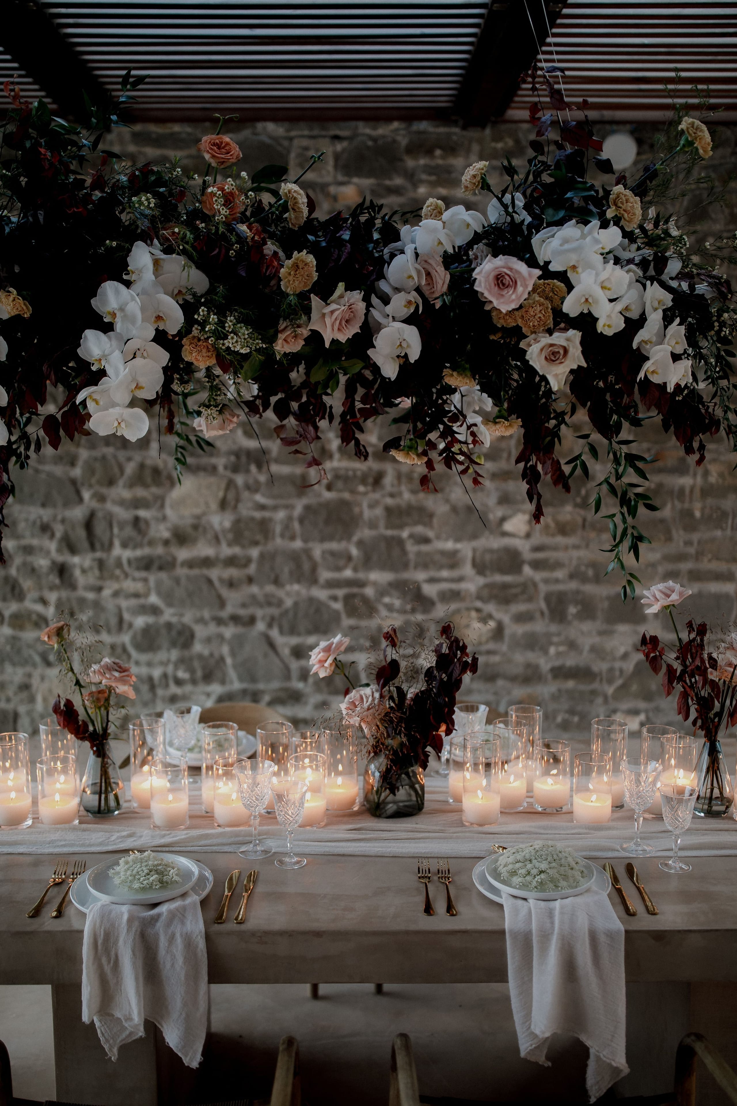 A suspended floral installation of white phalaenopsis orchids, blush and cream roses, apricot blooms and deep burgundy foliage hangs above a long wooden wedding table at Nuez Villas in Crete, Greece. Below, rows of pillar candles glow in glass hurricane holders, with small bud vases of dark and pink blooms set along a sheer runner, and the plates are dressed with white napkins. The old stone wall behind and the soft, low light create an intimate, romantic mood.