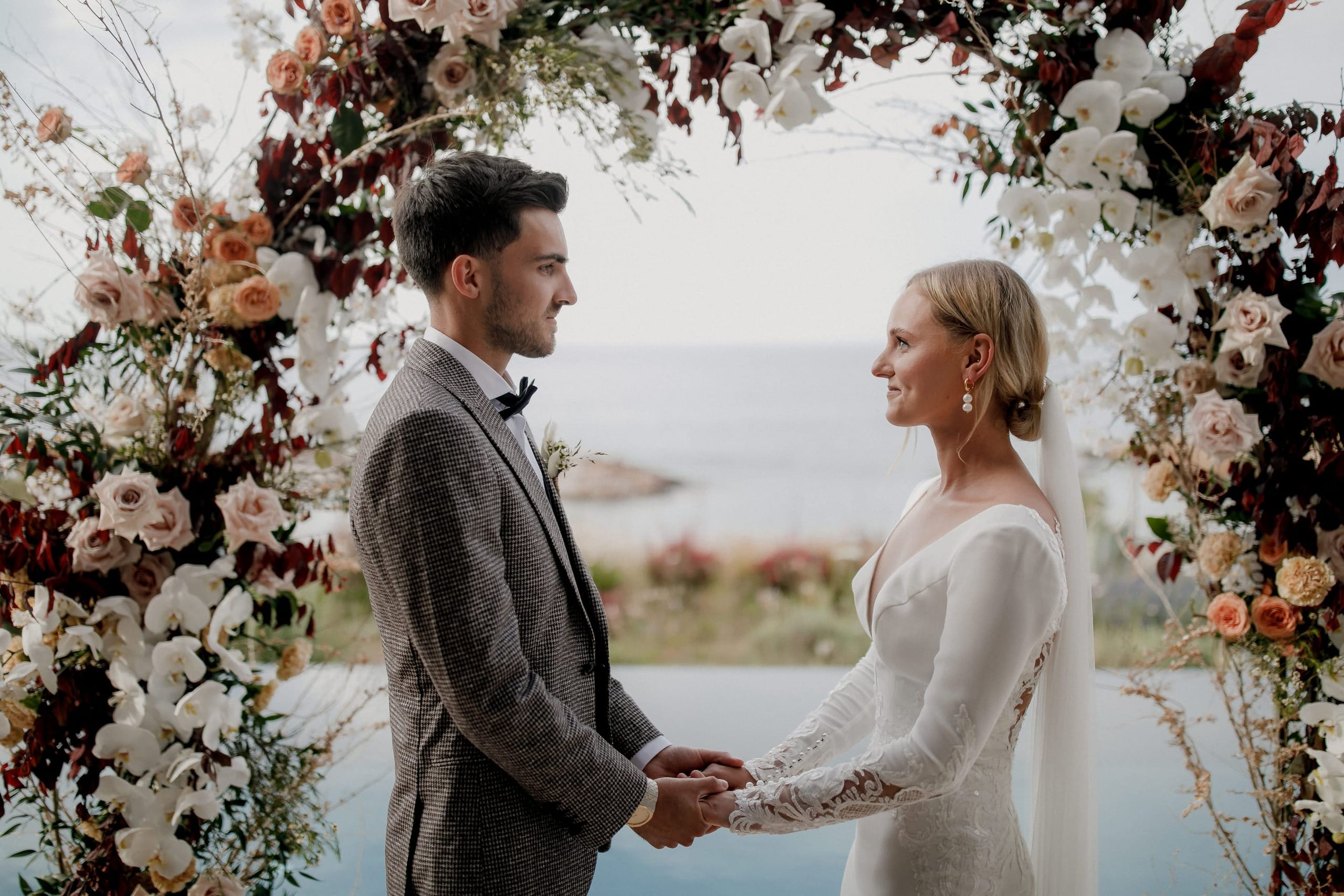A bride and groom stand hand in hand facing each other beneath a wedding arch at Nuez Villa in Greece, their gazes locked in a quiet moment. The arch is woven with white phalaenopsis orchids, peach-toned roses, deep burgundy foliage and wispy sprigs, and the sea glows pale behind them. He wears a grey houndstooth suit with a black bow tie, while she wears a long-sleeved white gown with lace detailing and a flowing veil, her hair swept into a low chignon.