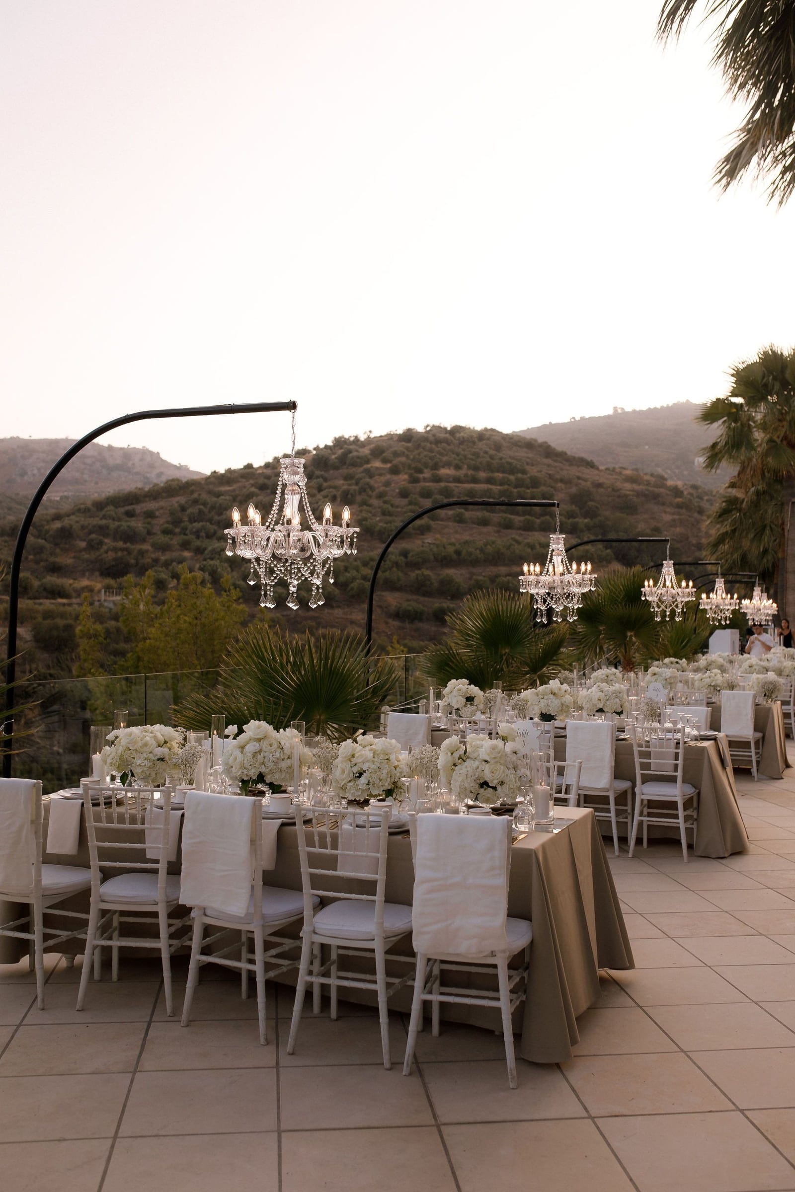 Long rows of wedding tables on a stone terrace at dusk, dressed in taupe linen with white chiavari chairs and white-draped chair backs. Low centrepieces of white roses, hydrangeas and baby's breath sit among tall candles, while crystal chandeliers hang from black arches above, and olive-covered hills and fan palms fill the view beyond the glass railing. This is a wedding reception setting in Greece.