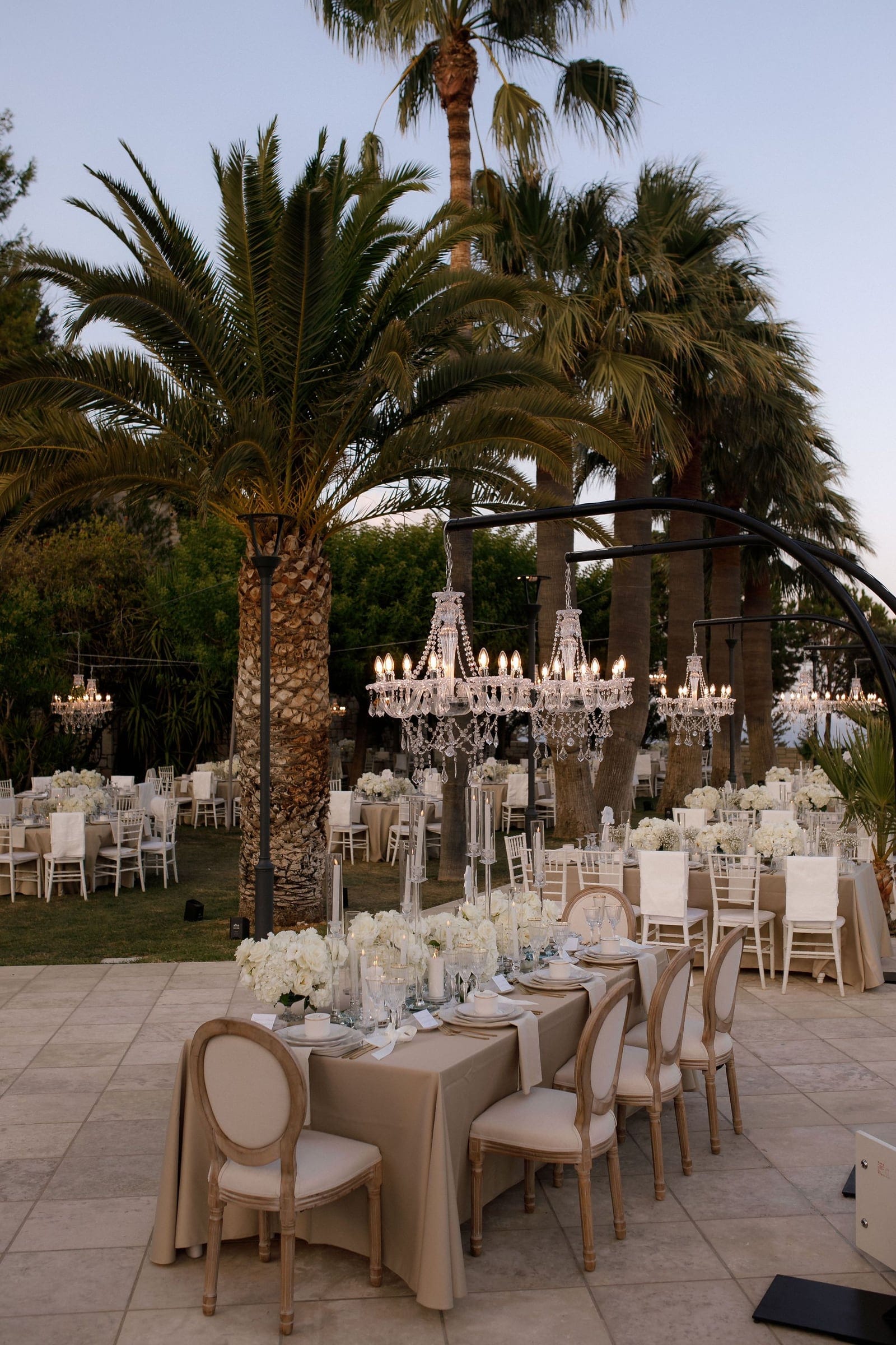 Crystal chandeliers hang lit at dusk above round tables dressed in white and cream, each centred with clusters of white roses and hydrangeas, in a garden at a wedding in Greece. Tall palm trees rise behind them against a pale evening sky, and a dark metal arbour frames the scene on the right. The white chairs and soft candlelight give the lawn a calm, elegant glow.