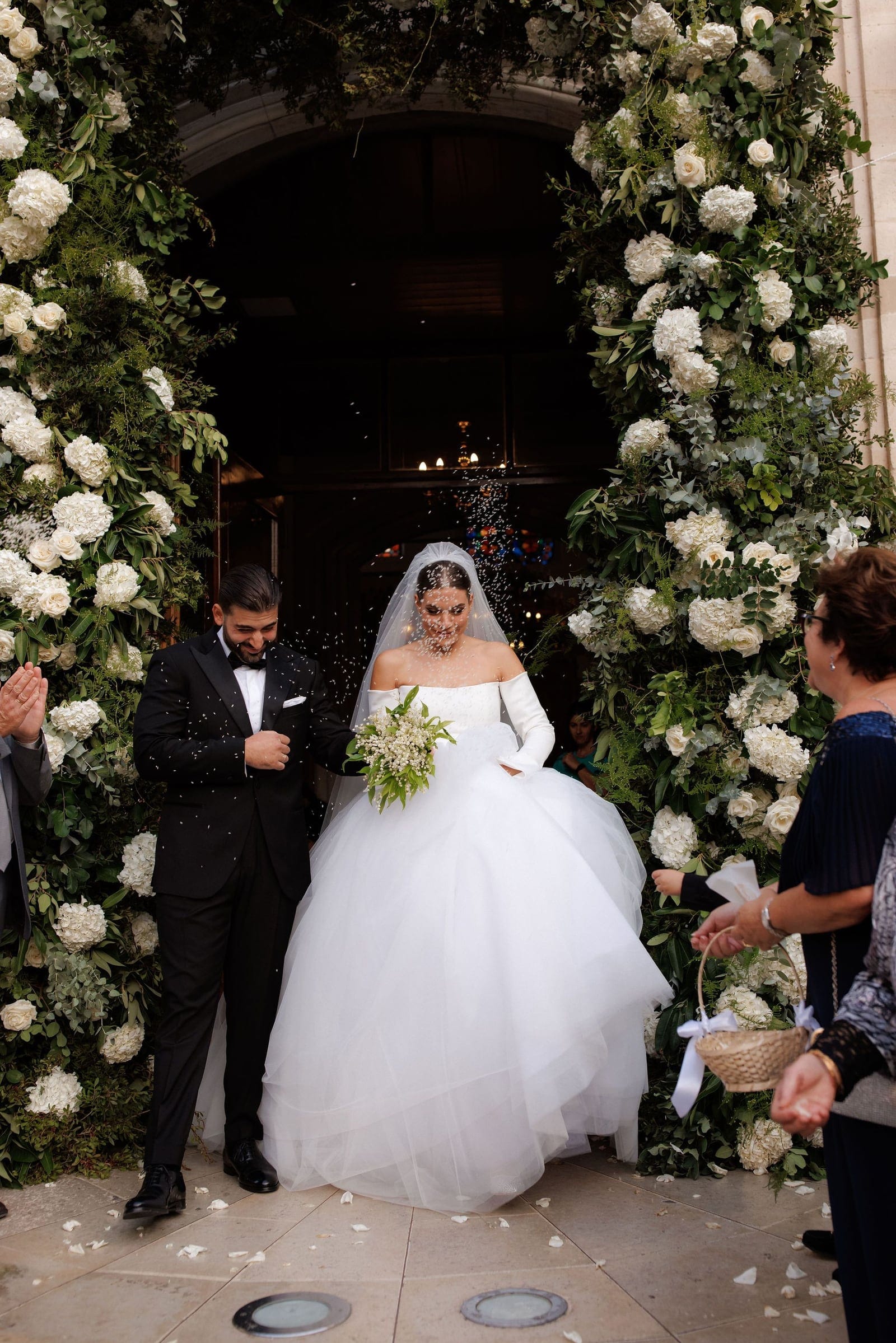 A bride and groom step out through a church doorway framed by tall arches of white hydrangeas, white roses and silver-green eucalyptus, all part of the wedding flowers for this Greece celebration. Confetti drifts through the air around them as they walk forward, the bride in a voluminous white ballgown with long gloves and a veil, holding a small bouquet of white blooms and greenery, while the groom in a black tuxedo smiles and looks down. Guests on the right watch and applaud, and the dim interior behind them glows with candlelight.