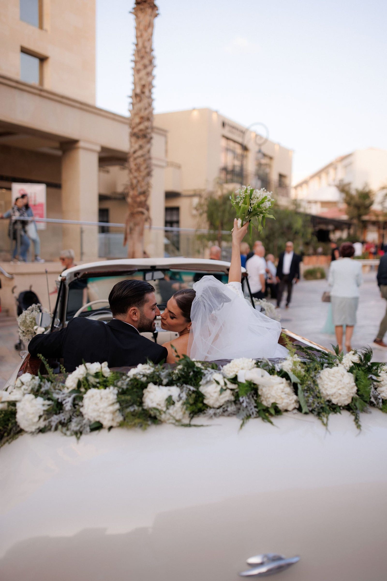 A bride in a white tulle veil and a dark-suited groom share a kiss in the back of a vintage white convertible, while she raises a small bouquet of white lily of the valley high in the air. The car is dressed with a garland of white hydrangeas and sprigs of silvery foliage, and guests stroll along a sunlit street behind them. These wedding car flowers were arranged by manfler in Greece.