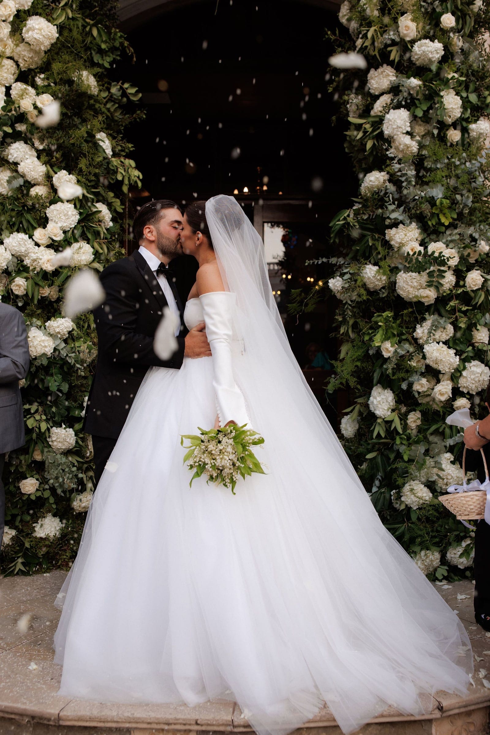 A bride and groom share a kiss beneath a tall doorway framed by lush arches of white hydrangeas, white garden roses and eucalyptus, while white petals drift through the air around them. The bride wears a voluminous white ball gown with off-the-shoulder long sleeves and a flowing veil, and holds a bouquet of lily of the valley wrapped in green foliage. The groom wears a black tuxedo and bow tie. Wedding flowers in Greece, with a warm, glowing interior behind them.