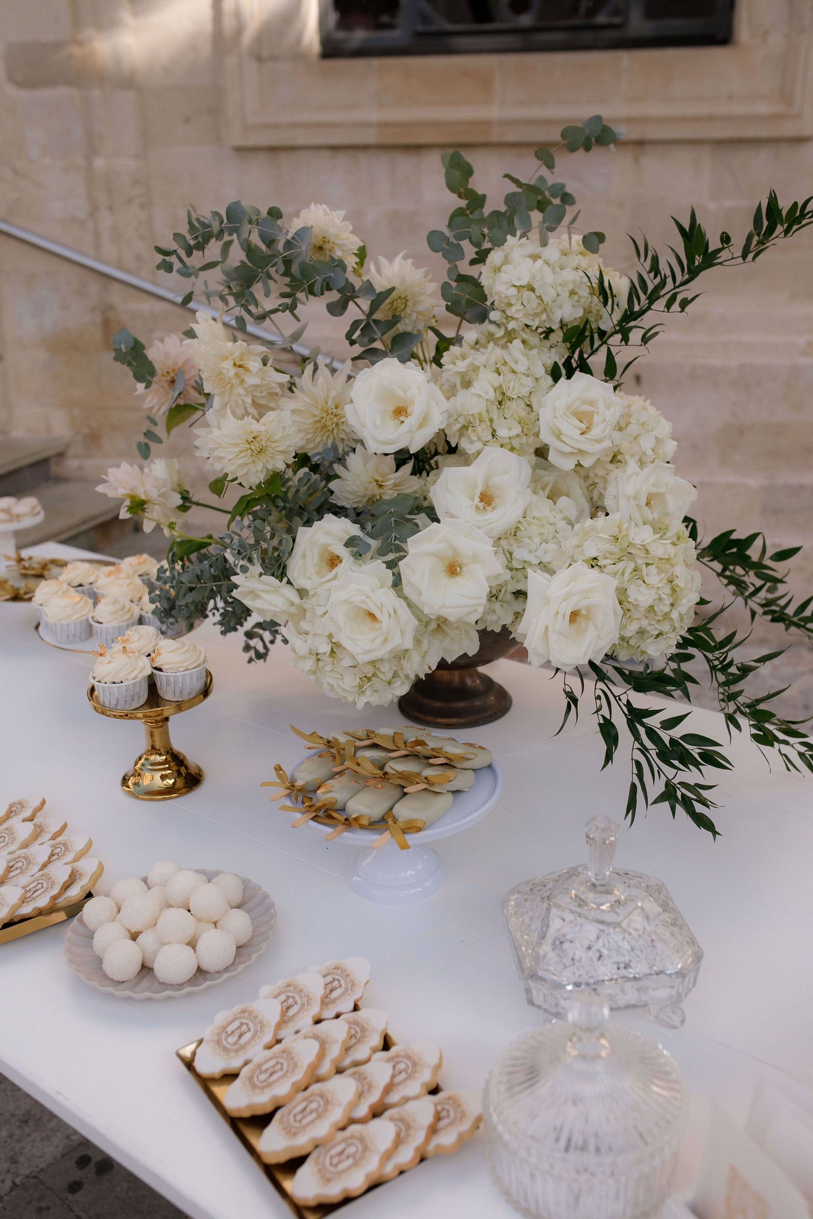 A tall, lush arrangement of cream garden roses, white hydrangeas and pale dahlias spills from a dark bronze pedestal urn, trailed with silvery eucalyptus and long green foliage. It sits on a white dessert table at a wedding in Greece, beside gold-rimmed cupcakes, glazed biscuits tied with gold ribbon and a cut-glass lidded dish, under soft natural light.
