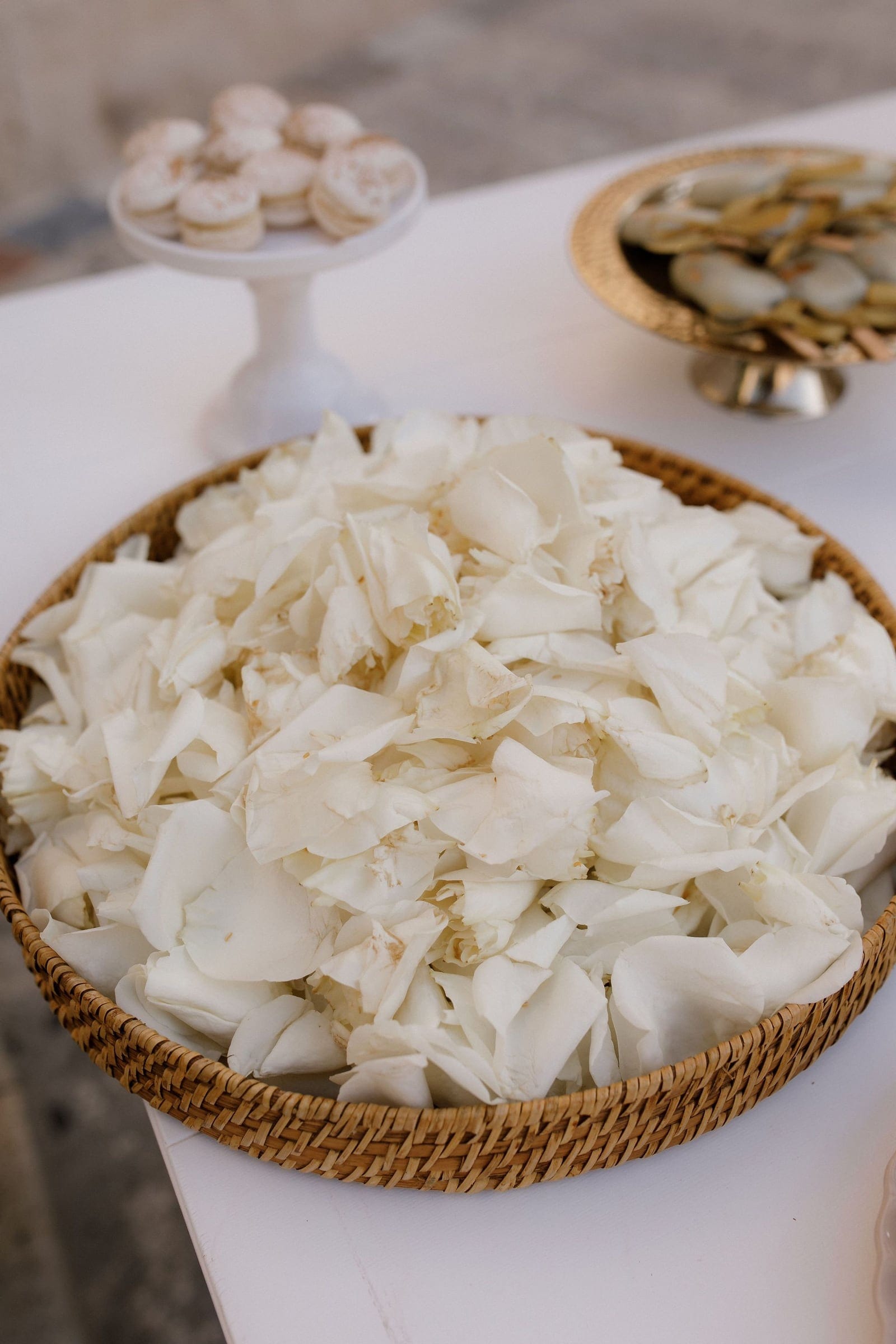 A woven wicker basket heaped with loose white rose petals, their creamy edges curling softly against one another, sits on a white tablecloth. Behind it, a gold-rimmed pedestal dish and a white candle or vase blur into the background, catching the soft daylight. These petals are wedding flowers from Manfler, ready to be scattered for a ceremony in Greece.