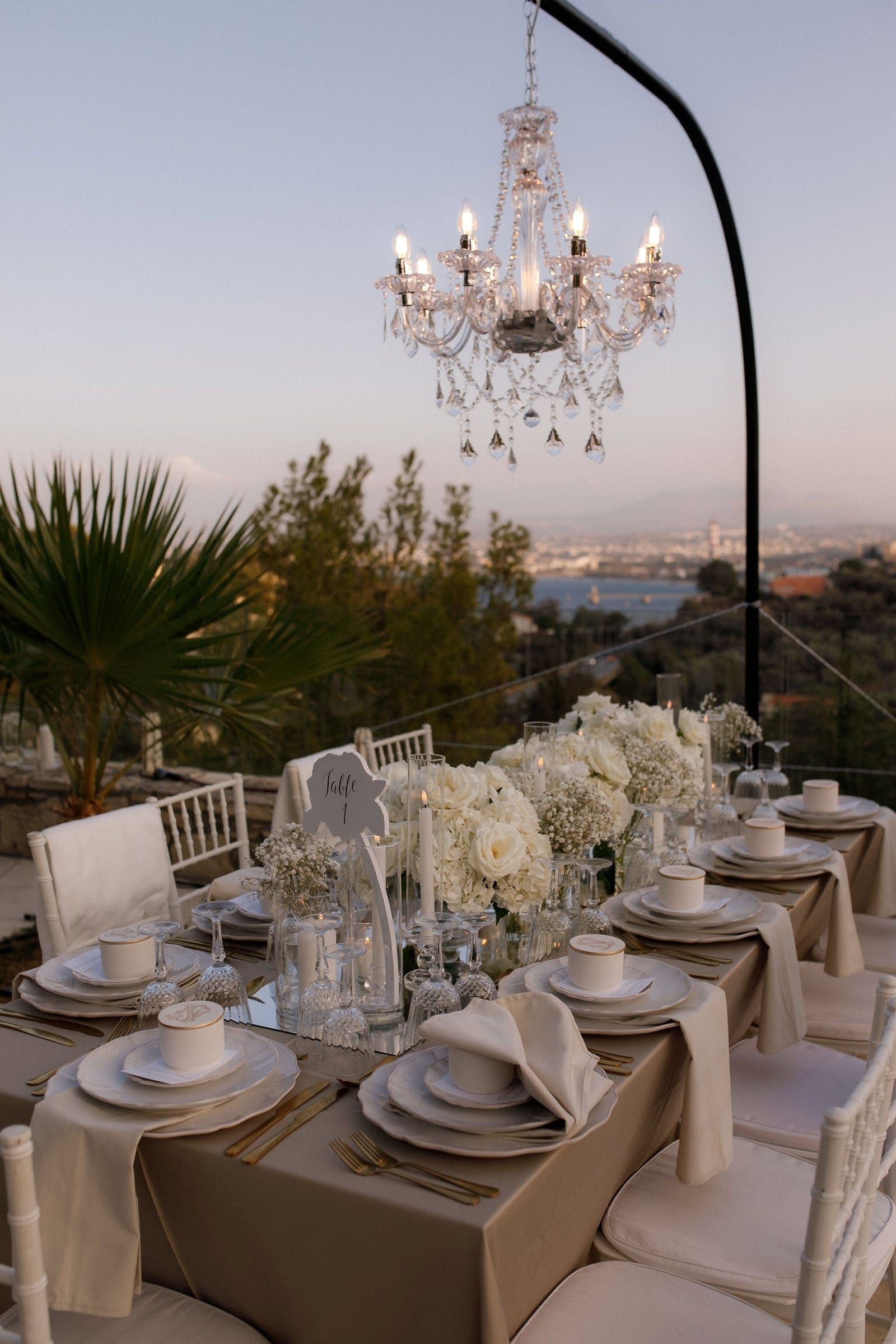 A long wedding dinner table outdoors at dusk, dressed in cream linens with gold-rimmed white plates, crystal glasses and gold cutlery. The centrepiece is a lush arrangement of white garden roses, white hydrangeas and baby's breath in glass vases, with tall white candles flickering beside it and a grey "Table 1" sign standing among them. Above hangs a crystal chandelier, and beyond the glass canopy a hazy sea and town glow in the evening light, with a palm and green trees at the edge. This is a wedding table setting by manfler, a florist in Greece.