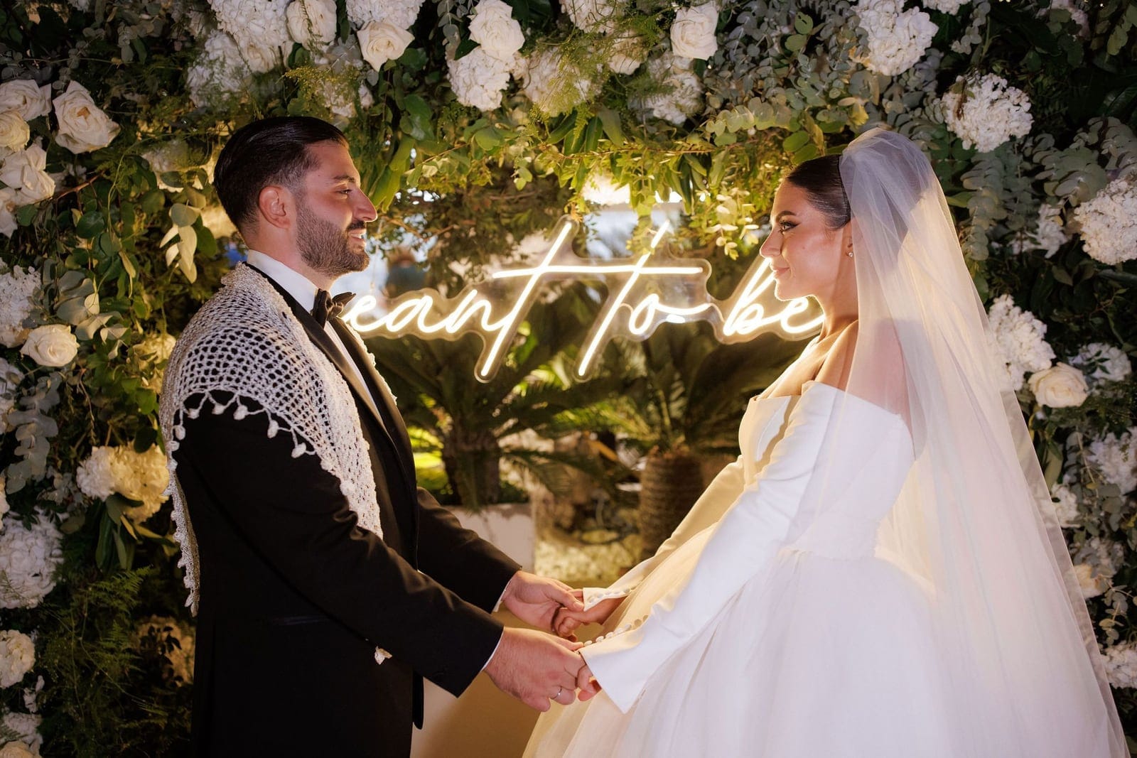 A bride and groom stand facing each other in profile, holding hands in front of a floral installation of white garden roses, hydrangeas and green eucalyptus. Behind them a glowing white neon sign reads "meant to be," with palm fronds softly blurred in the evening light. He wears a black tuxedo and a white crocheted shawl over his shoulders, and she wears a white gown with long sleeves and a veil. This wedding flower display is in Greece.