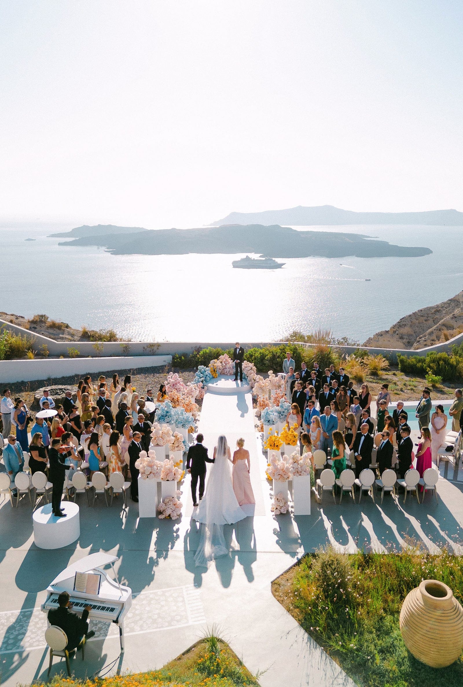Seen from above, a bride in a long veil and a groom in a dark suit walk hand in hand down a white aisle toward a raised white platform, while a bridesmaid in a blush gown follows beside them. The aisle is lined with pastel arrangements of garden roses, hydrangeas and blue and yellow blooms on white pedestals, in a wedding ceremony at Eros Santorini in Greece, with the sea sparkling beyond the terrace.