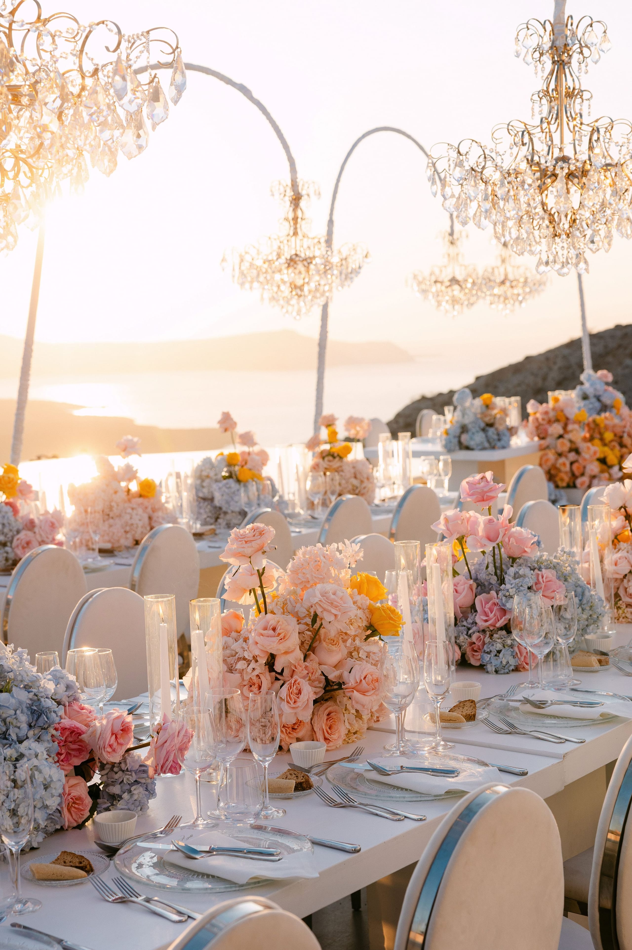 A long wedding dinner table at sunset, set on a terrace at Eros Santorini in Greece, with the caldera sea glowing gold behind it. Low centrepieces of blush garden roses, pale peach hydrangeas and sprigs of yellow roses spill over the white cloth, interspersed with powder-blue hydrangeas. Tall taper candles stand in glass cylinders between clear glasses and cream chairs, and the warm light gives the whole scene a soft, romantic glow.