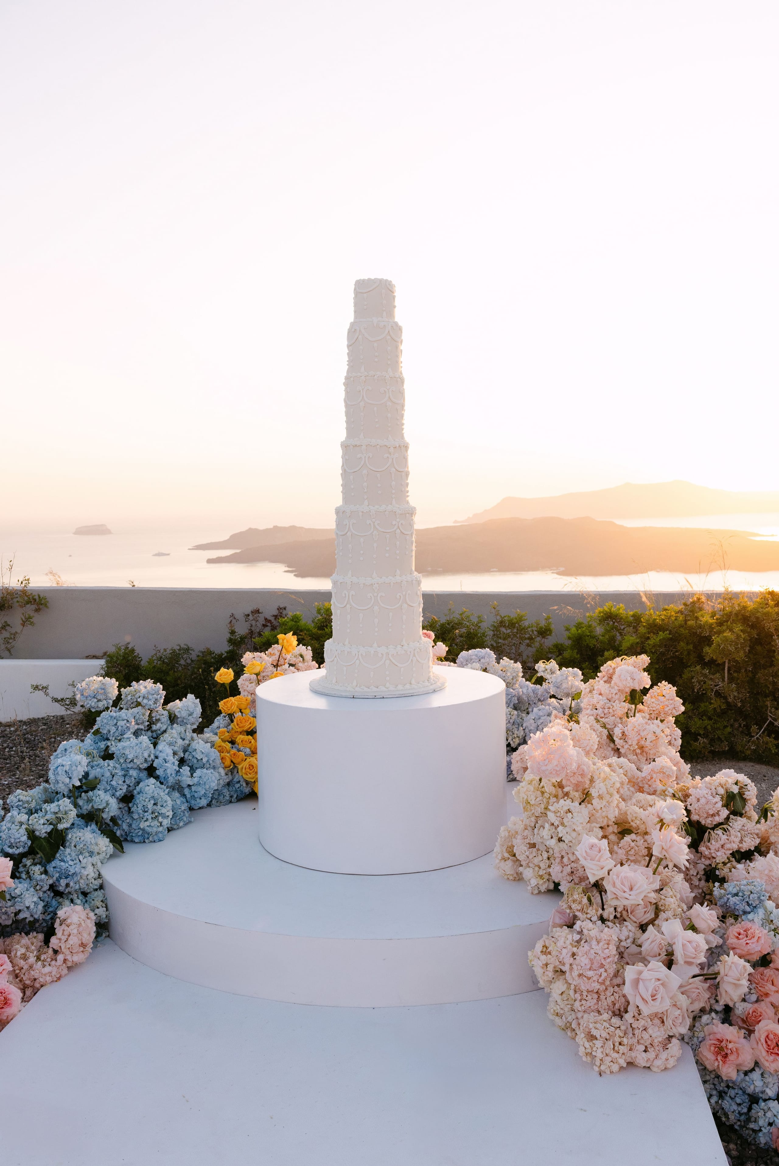 A tall, tiered white wedding cake, piped with delicate scrollwork and beading, stands on a white circular pedestal at Eros Santorini in Greece, framed by drifts of pale blue hydrangeas, blush roses and cream hydrangeas, with small yellow roses tucked in. The warm, golden sunset light softens the scene against a hazy view of the Aegean and distant islands.