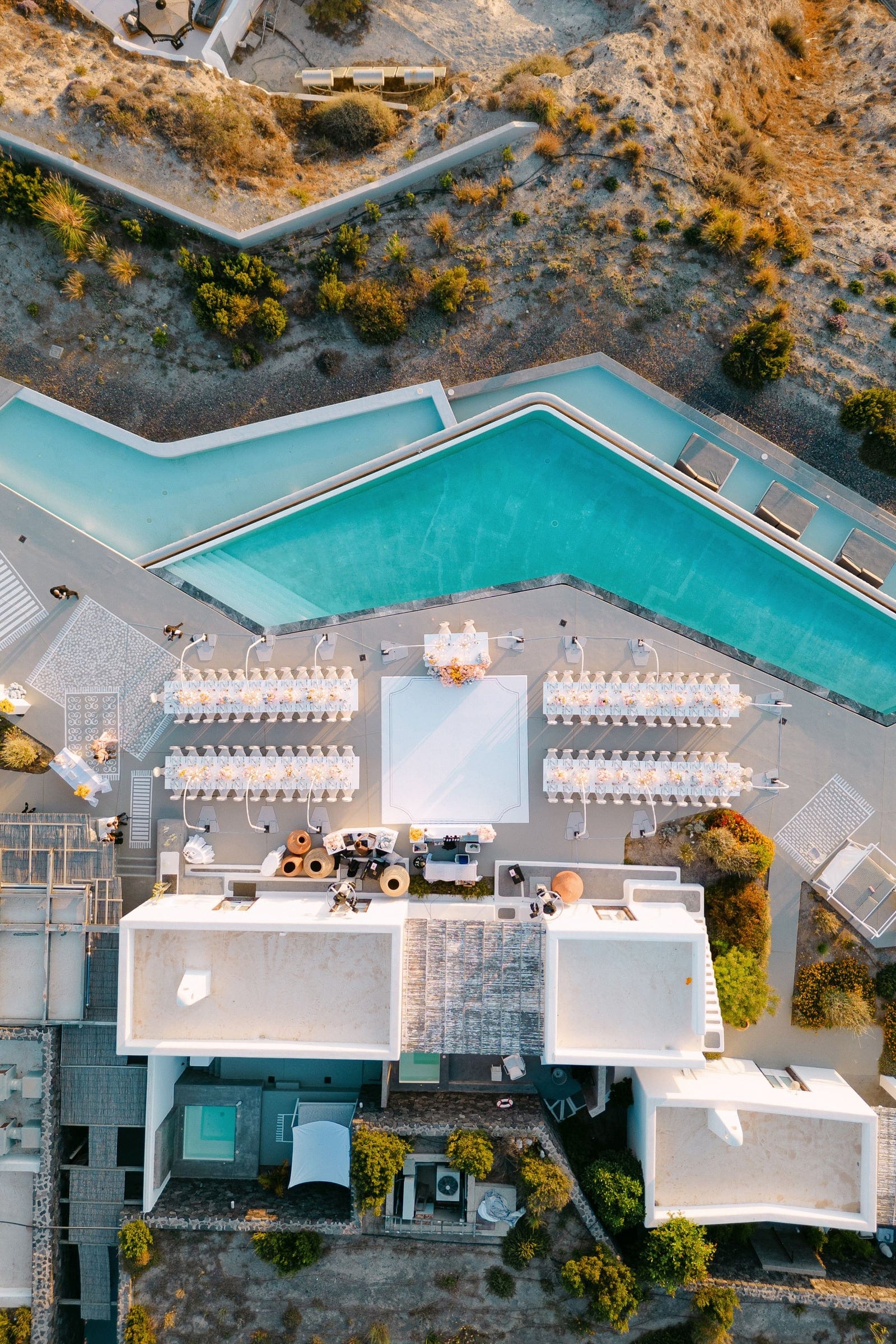 An overhead aerial view of a wedding reception at Eros Santorini in Greece, showing four long banquet tables dressed in white linen and lined with low arrangements of cream and pale peach blooms, likely roses and orchids. A large white dance floor sits at the centre beside a floral centrepiece, all beside a turquoise infinity pool and white-washed villa rooftops, with a sunlit terrace and a strip of golden-orange shrubs along the edge.