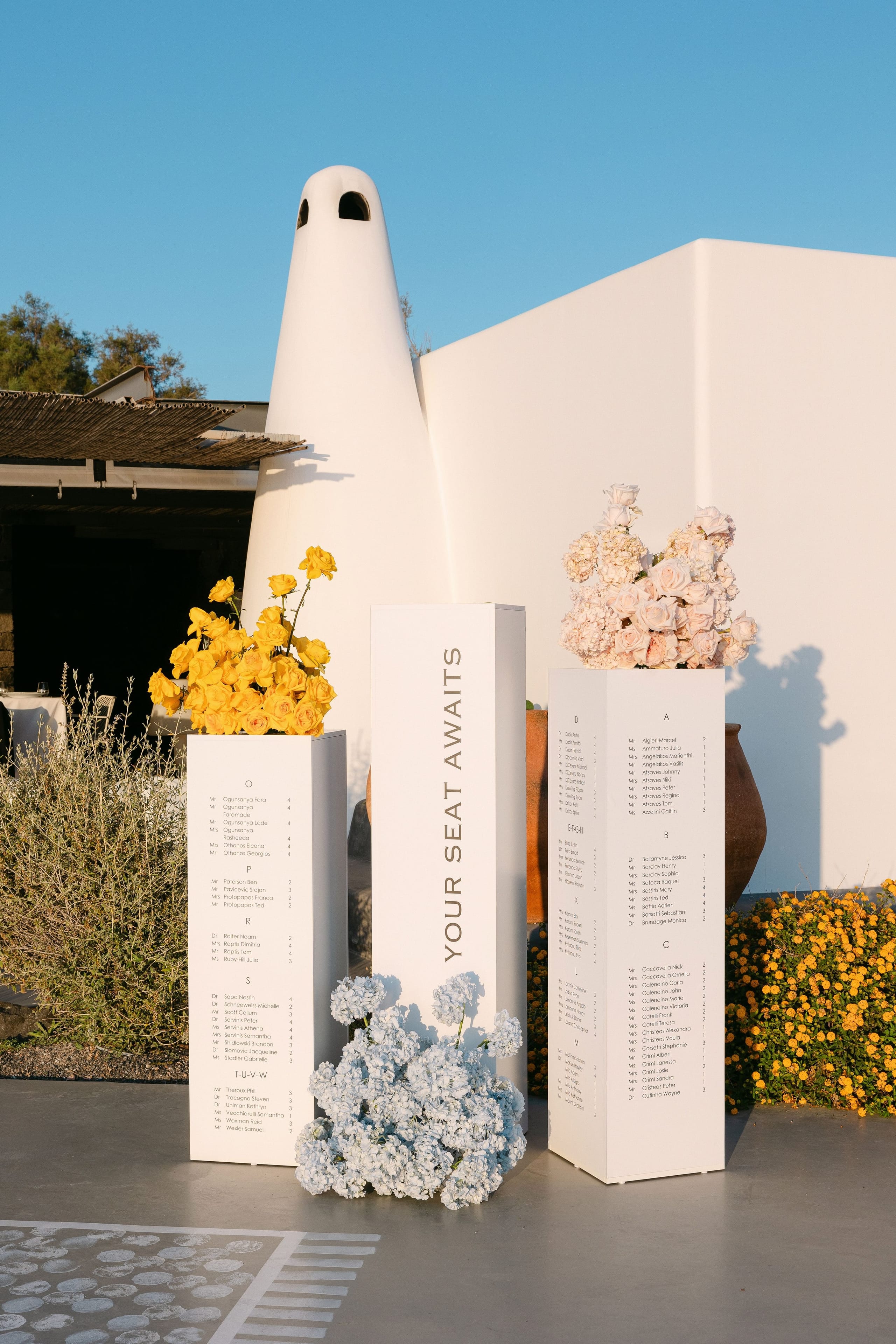 Three tall white plinths stand on a grey terrace at Eros Santorini in Greece, each printed with alphabetised wedding seating names, with the central one reading "Your seat awaits" in grey letters. Bright yellow roses spill from the left plinth, pale cream roses and hydrangea from the right, and a low cascade of pale blue hydrangeas sits at the base of the middle one, all against a whitewashed wall in warm evening light.