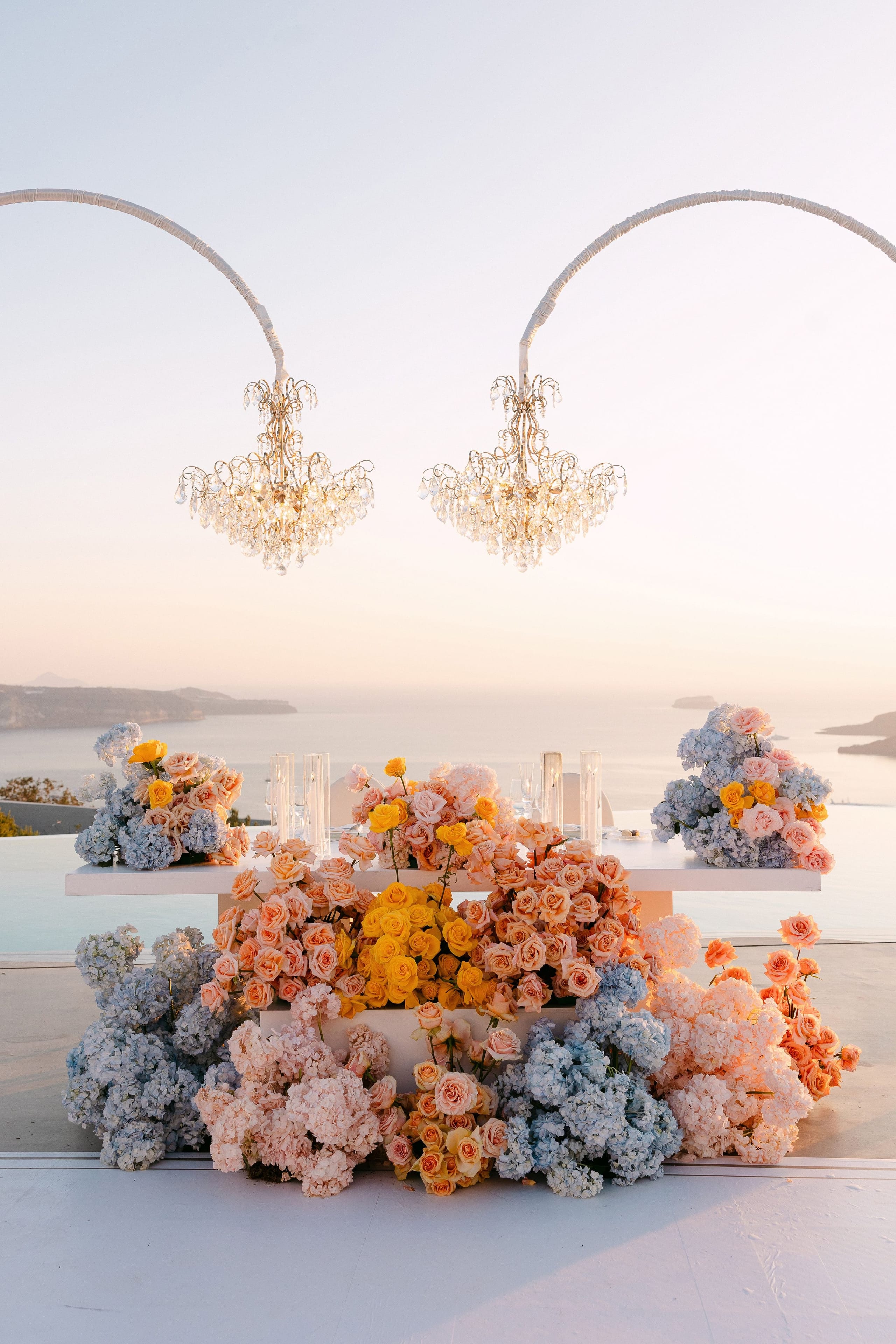 A long white sweetheart table on a clifftop terrace at Eros Santorini, Greece, is dressed in cascading wedding flowers that shade from pale blue to peach to golden yellow. Tumbling mounds of blue hydrangea and blush hortensia spill across the floor and the table's base, while peach garden roses and yellow roses rise in lush arrangements, with orange blooms trailing at the right. The soft pink light of sunset lies over the calm sea and distant islands behind.
