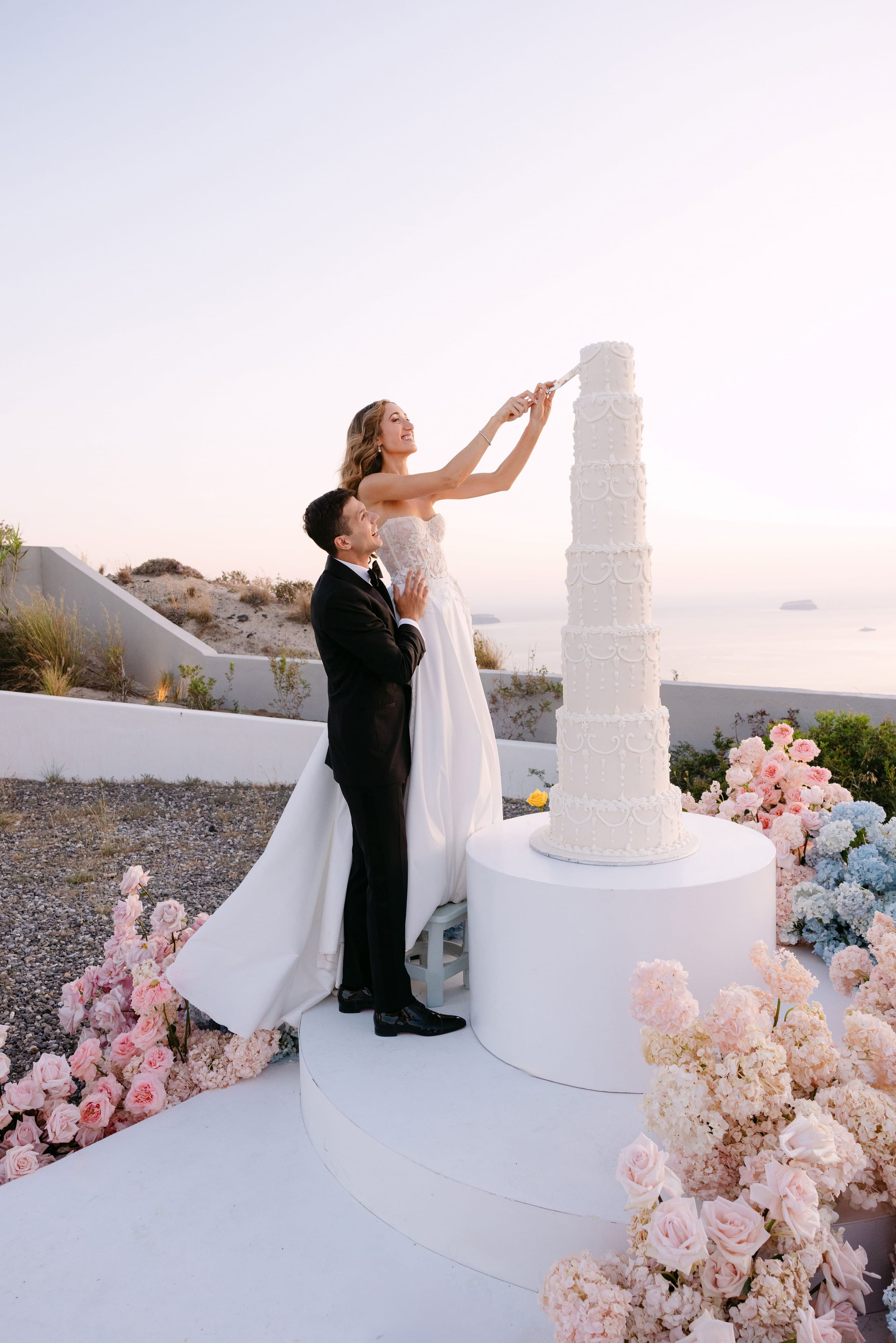 A bride in a strapless white gown and a groom in a black tuxedo reach up together to cut a tall, tiered white cake with piped scrollwork, set on a round white pedestal on a terrace at Eros Santorini in Greece. Pale pink garden roses, cream hydrangea-like blooms and soft blue hydrangeas spill across the ground around them, and the sea and a distant islet glow under a pale pink sunset sky. These are wedding flowers in Greece.