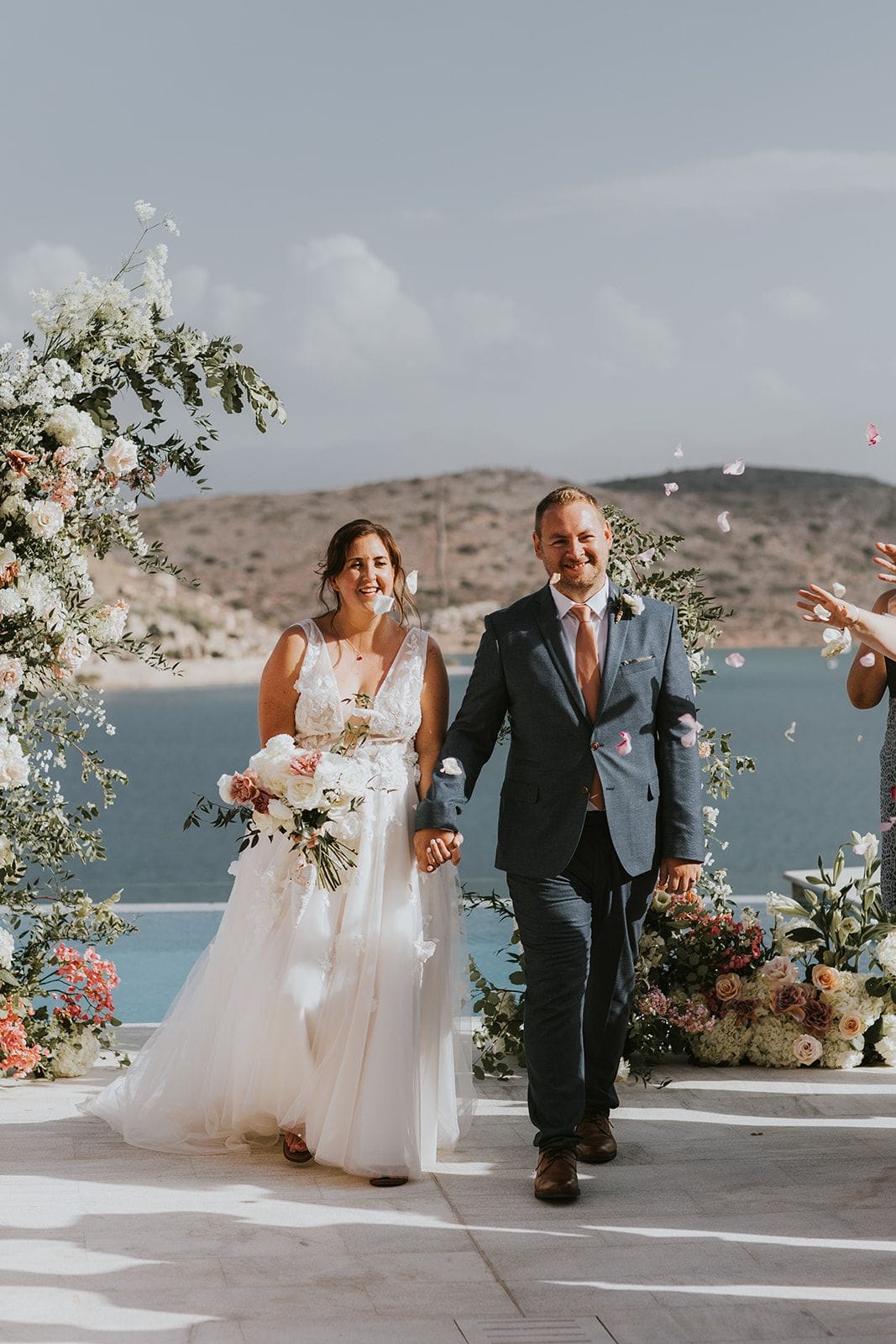 A bride and groom walk hand in hand past a floral arch of white roses, eucalyptus and trailing greenery at Villa Gina in Greece, while petals drift down around them. She carries a cascading bridal bouquet of white garden roses, blush and dusty-rose blooms, and wears a lace-bodiced tulle gown; he wears a slate-blue suit with a peach tie. Behind them, a calm blue bay and dry, rocky hills stretch out in soft afternoon light, and pale floral arrangements line the terrace.