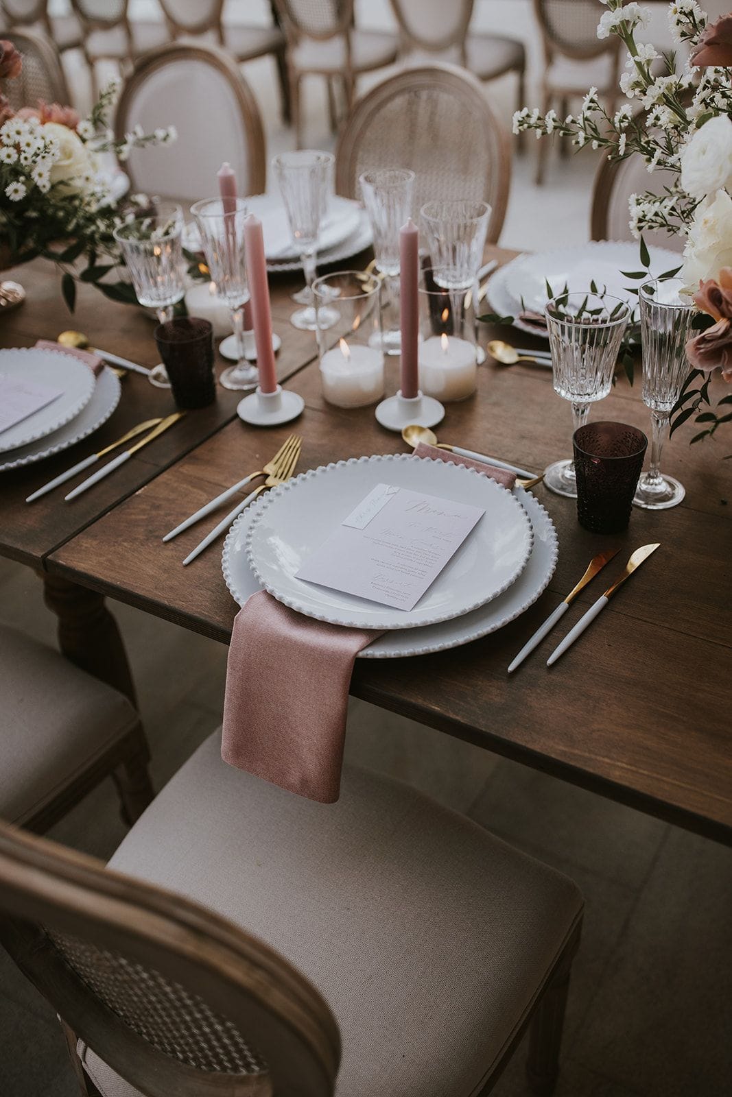 A wooden wedding table set for a reception at Villa Gina in Schisma Eloundas, Crete, Greece. A white beaded-rim plate holds a printed menu card, with a dusty-pink napkin draped beneath and gold-handled cutlery on each side. Pale pink taper candles stand among clear glasses, dark plum tumblers and small white pillar candles, while cream roses and white sprays of blossom fill the arrangements at the far edges.