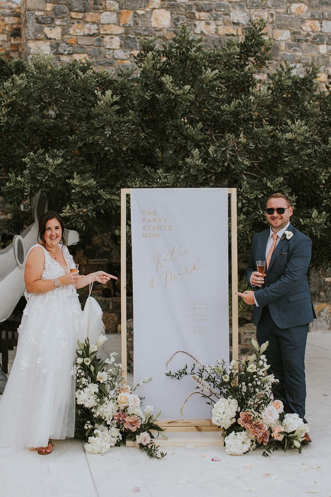 A bride in a white tulle gown with floral appliqué and a groom in a navy suit with a blush tie pose beside a tall wooden welcome sign reading "The Party Starts Now" and the couple's names, both raising champagne flutes and pointing at it. Ivory and blush arrangements of roses, hydrangeas and lilies spill across the stone base, with olive-green foliage behind, at Villa Gina in Schisma Eloundas, Crete, Greece, for a wedding.