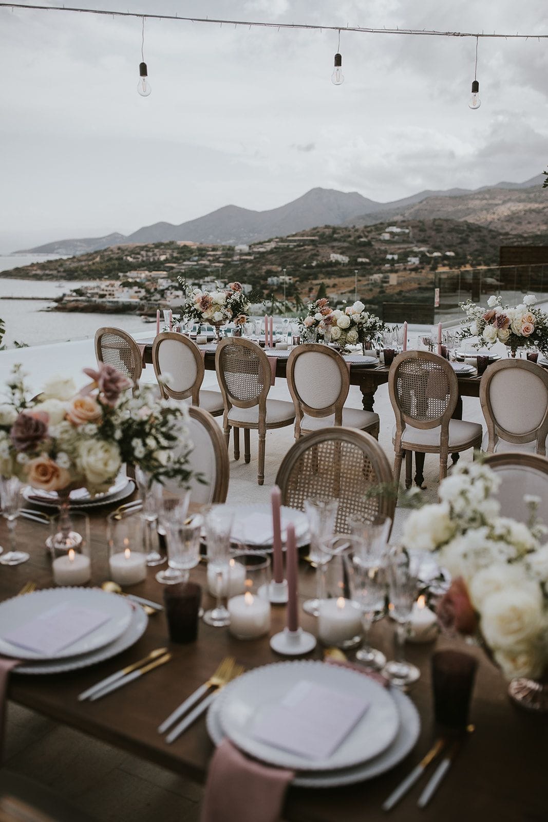 A long wooden wedding table set on a terrace at Villa Gina in Schisma Eloundas, Crete, Greece, with cane-backed chairs and low centrepieces of cream and blush roses, peach and dusty mauve blooms, and white flowers. Pink taper candles and small glass votives run along the table, and strings of bare bulbs hang overhead against a soft, overcast sky. Beyond the terrace, the sea and a village on the shore sit below dark hills.