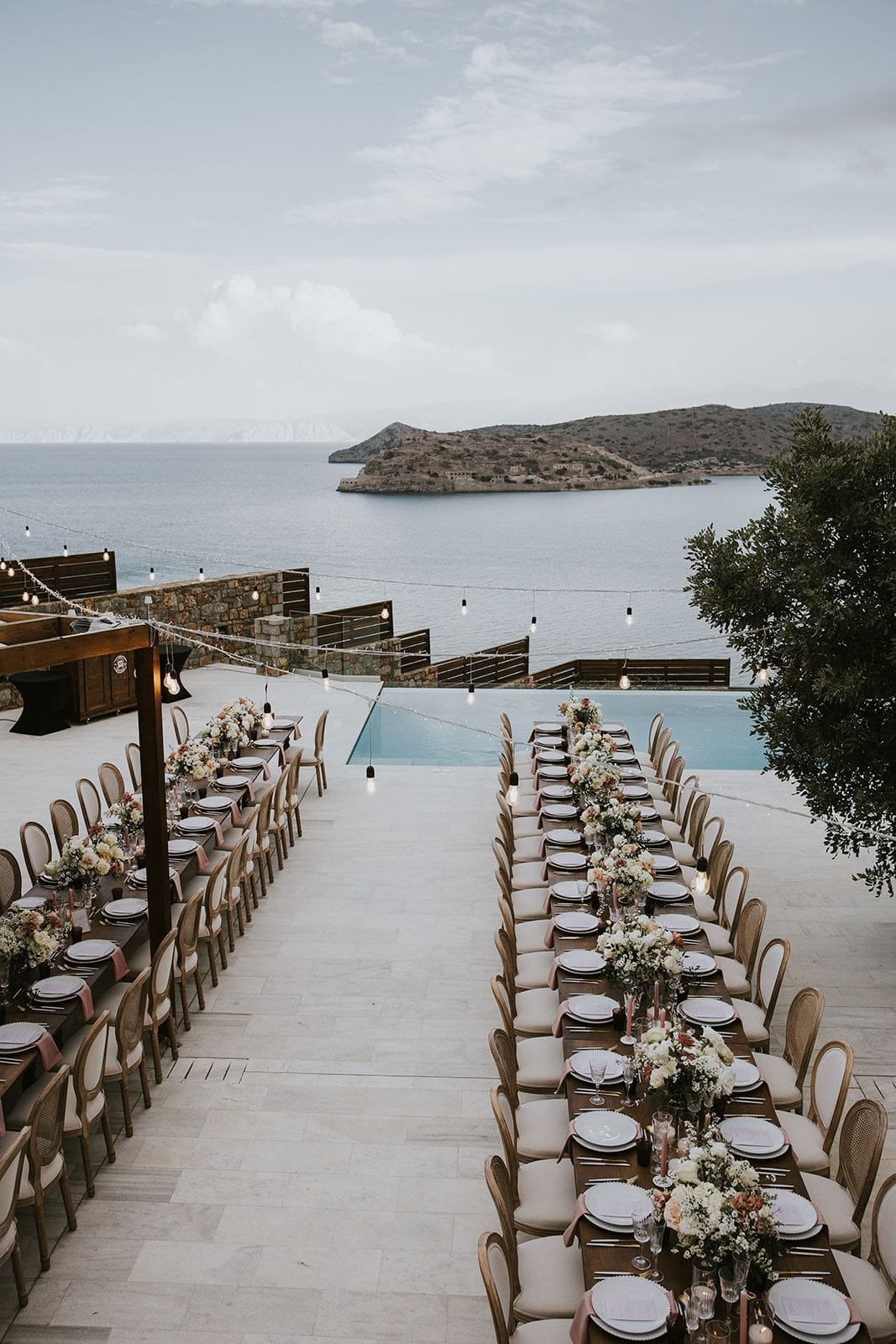 Long wooden banquet tables run in two rows across a pale stone terrace at Villa Gina in Schisma Eloundas, Crete, Greece, each laid with white plates, pink napkins and low arrangements of cream and blush blooms. Strings of festoon bulbs sag above the tables toward a sparkling infinity pool, beyond which the calm sea and a rocky island fade into a soft, hazy evening sky. It is a wedding table setting, with the flowers arranged for a sunset dinner by the water.