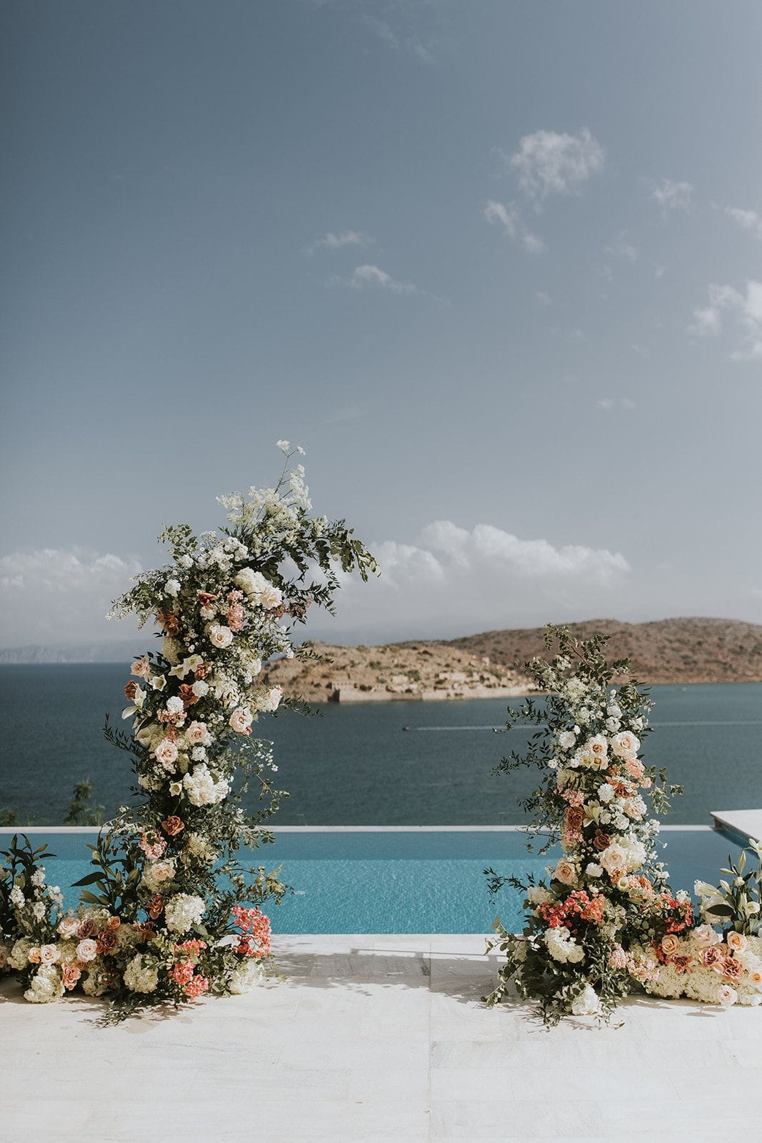 Two tall wedding arches framed in cream roses, white hydrangeas, pale peach blooms, coral flowers and trailing green foliage stand on a white stone terrace beside an infinity pool at Villa Gina in Schisma Eloundas, Crete, Greece. Behind them the blue sea stretches to a dry, rocky island under a pale sky with soft clouds. The mood is airy, romantic and sunlit, a ceremony setting for a wedding in Greece.