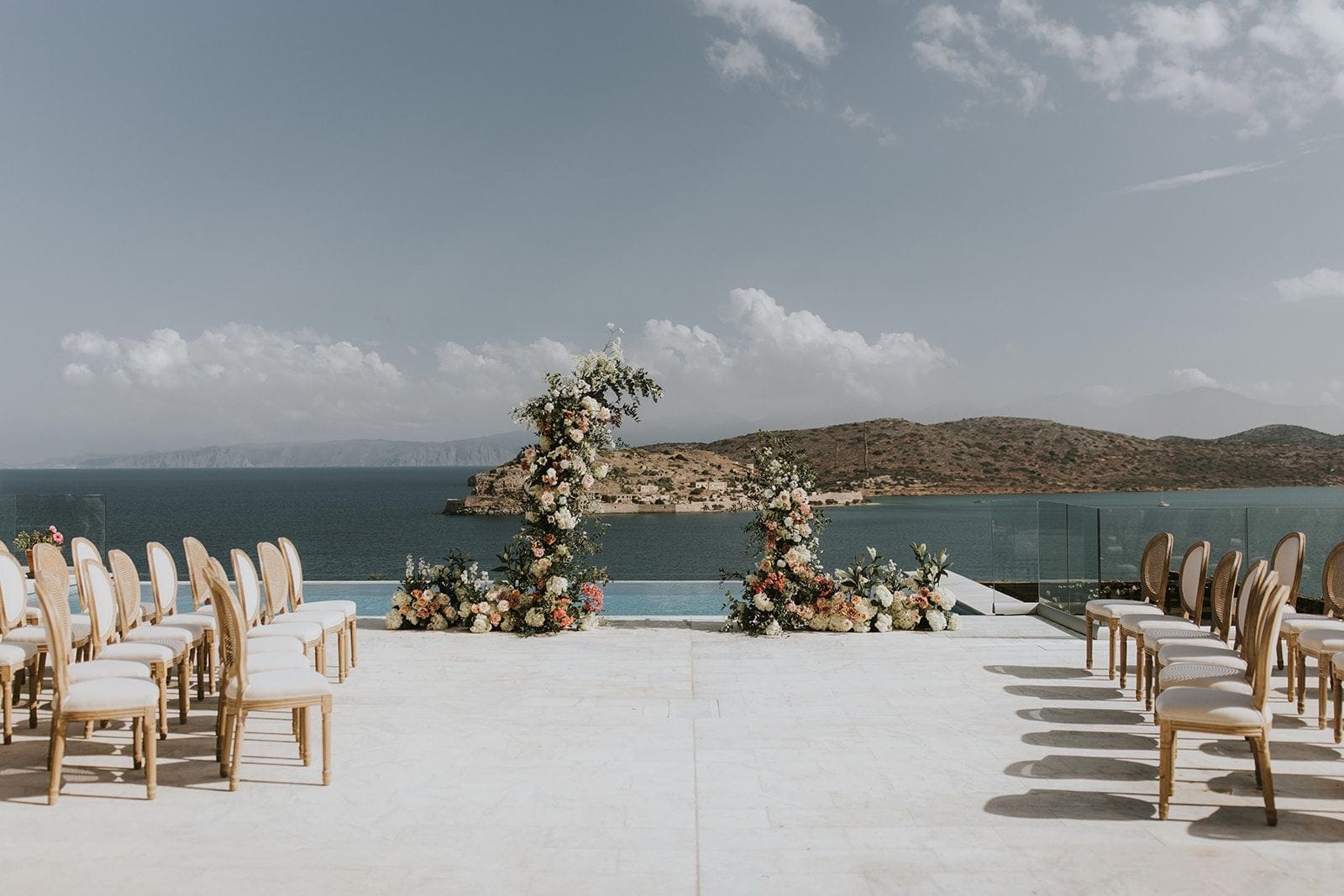 Two tall floral arches of white, cream and blush blooms, with greenery trailing up their sides, stand at the far edge of a pale stone terrace beside an infinity pool. Rows of cream-cushioned wooden chairs line both sides of an aisle that leads to them, with the calm blue sea and a brown, scrub-covered headland beyond. These wedding ceremony flowers at Villa Gina in Schisma Eloundas, Crete, Greece, are set under a pale, hazy sky.