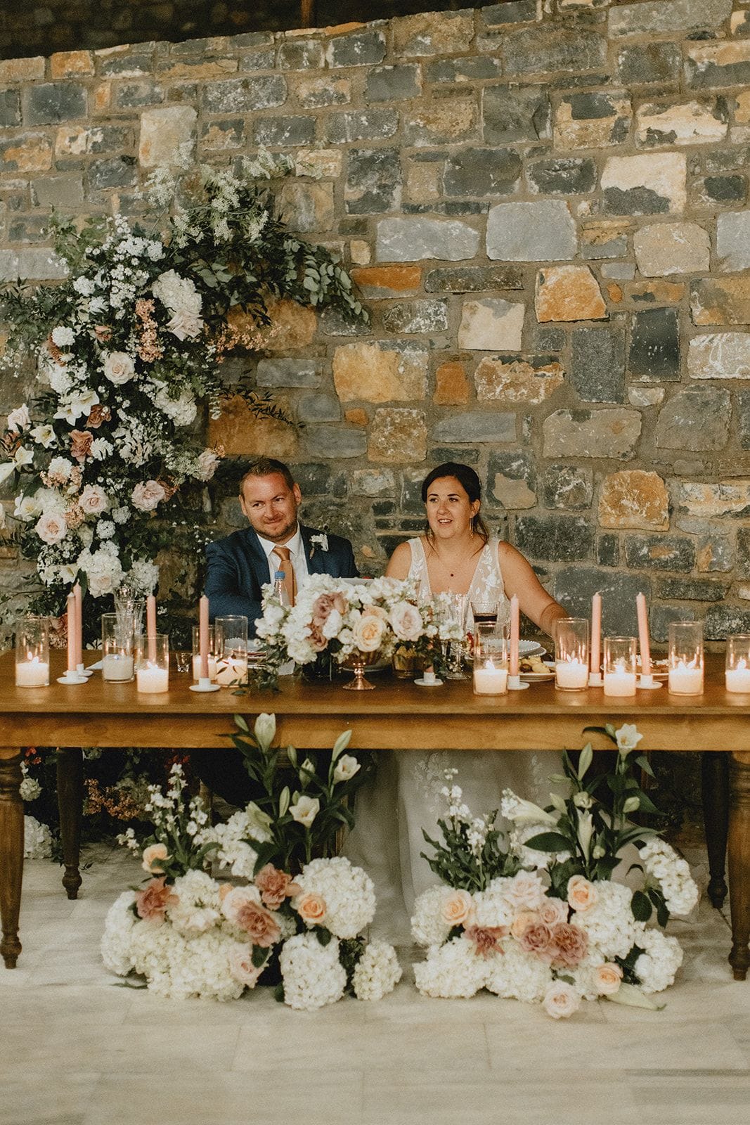 A bride and groom sit smiling behind a long wooden head table at Villa Gina in Schisma Eloundas, Crete, Greece, against a rough stone wall. Cascading white and blush flowers, including roses, hydrangeas and eucalyptus, rise up beside them, while pillar candles in glass hurricanes glow along the tabletop. Low clusters of white hydrangeas and peach roses spill across the pale floor below, and the groom wears a navy suit with a rust tie. This is a wedding flower setup in a warm, candlelit room.