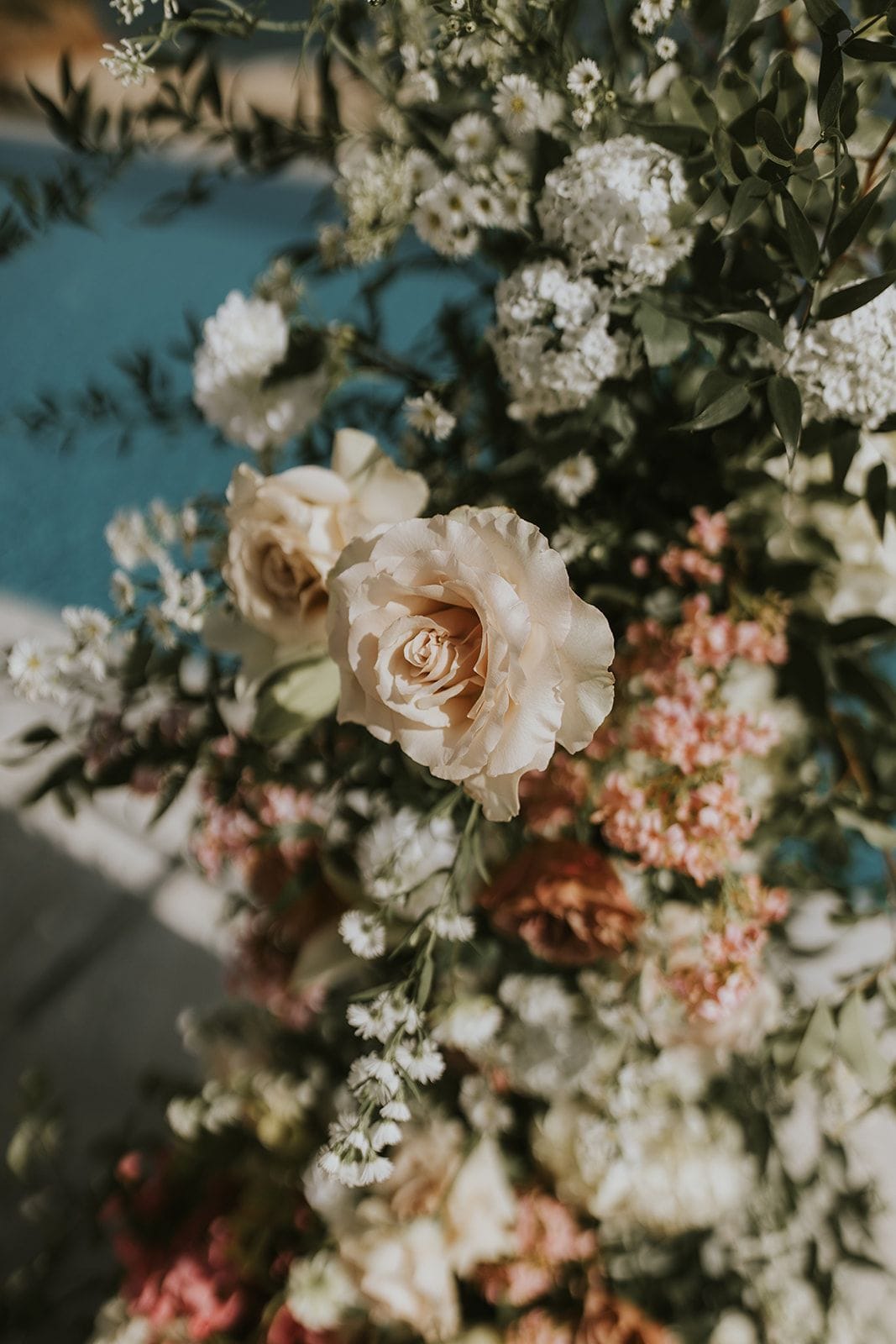 Cream and blush garden roses lead a lush arrangement of white gypsophila, pale pink blooms and trailing green foliage, with the blue of a pool softly blurred behind. Warm sunlight falls across the petals, giving the scene a soft, romantic glow. This is a wedding floral installation at Villa Gina in Schisma Eloundas, Crete, Greece.