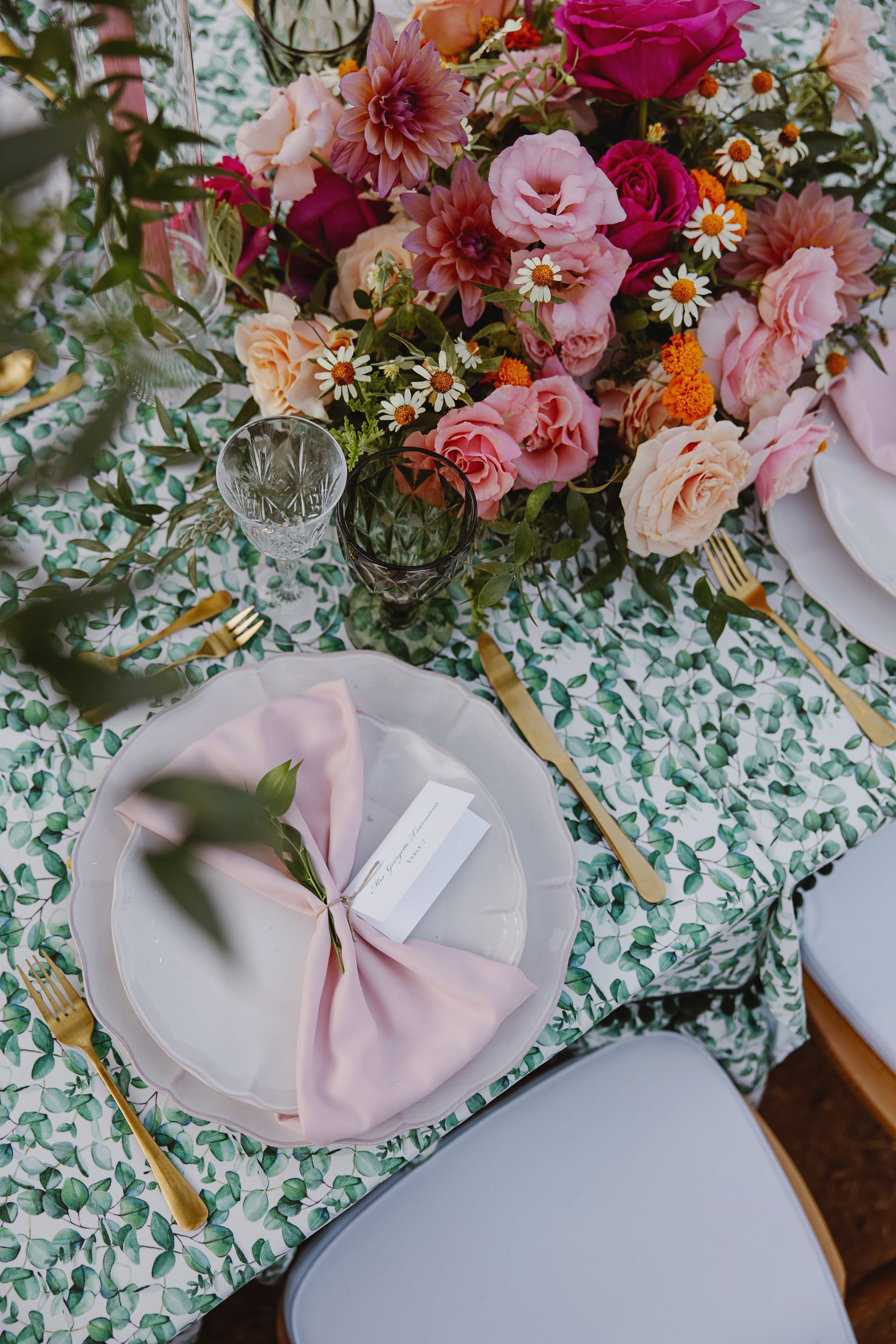 A top-down view of a wedding table set on a leaf-patterned cloth, with a lush centrepiece of pink garden roses, peach roses, dahlias, white daisies and orange marigolds. Gold cutlery, crystal and smoky green glasses, and a blush napkin on a white plate with a name card frame the arrangement, in Greece.