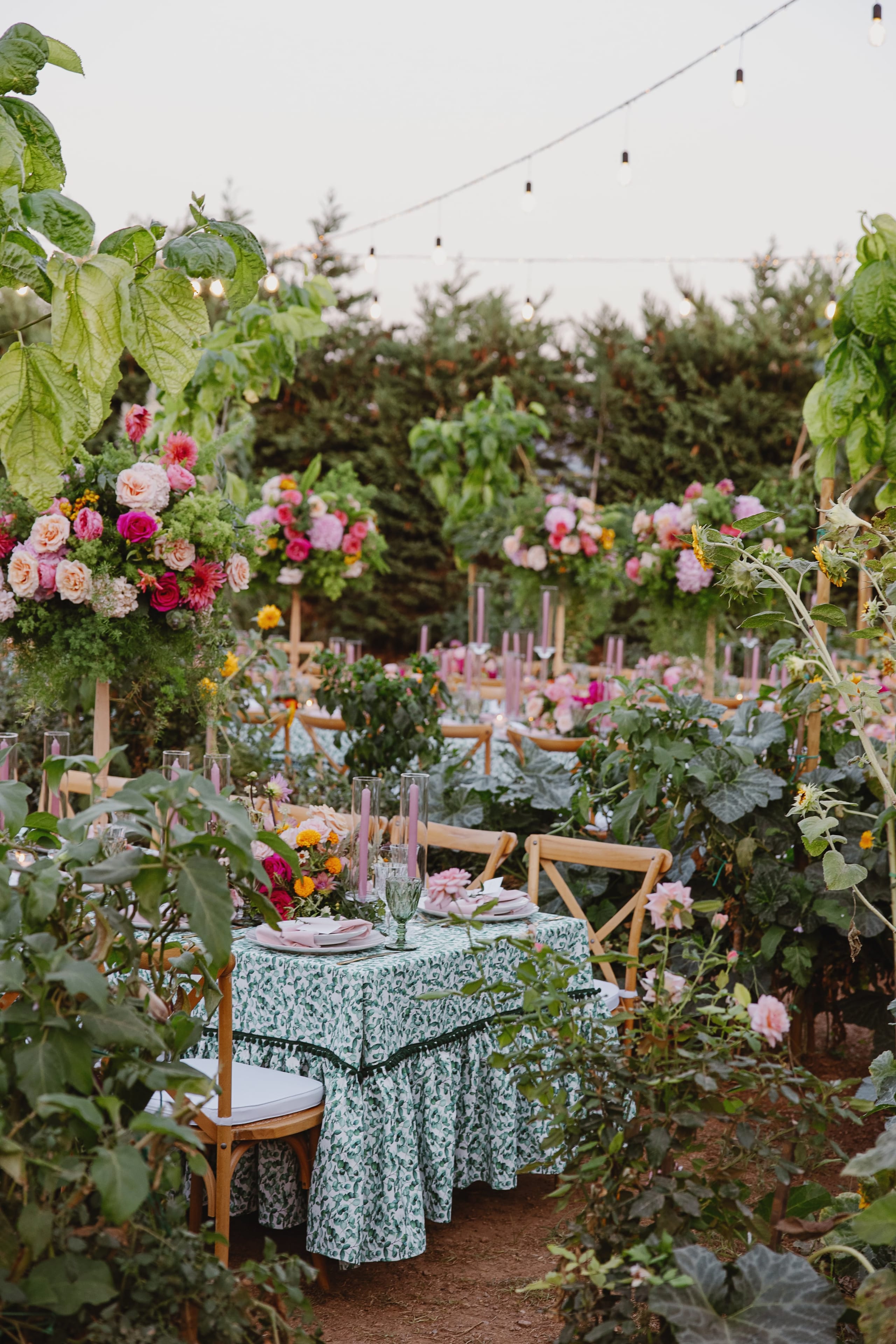 A wedding table setting in a lush garden in Greece, with a green and white patterned cloth, pink plates and lilac candles in glass hurricanes. Behind it, tall floral arrangements of pink roses, peonies and dahlias on wooden stands rise among leafy green plants and sunflowers, with rows of cross-back chairs stretching into the greenery.