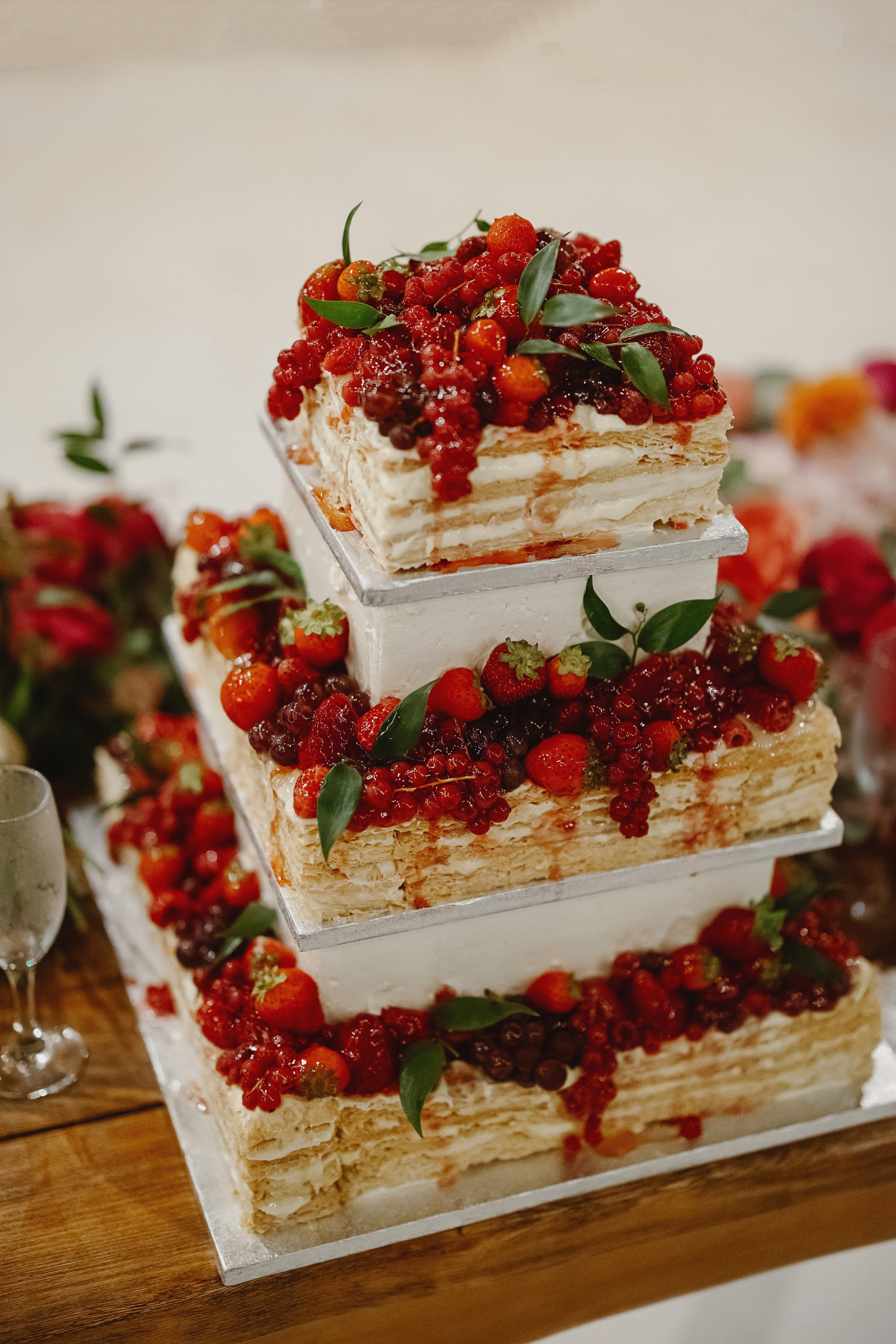 A three-tiered square wedding cake with layers of pale crêpe-like sponge and cream, each tier set on a silver board and crowned with glossy red currants, strawberries, dark blackcurrants and small green leaves. Blurred red and orange flowers sit in the background, and a glass of sparkling wine stands at the left edge of this wedding reception in Greece.