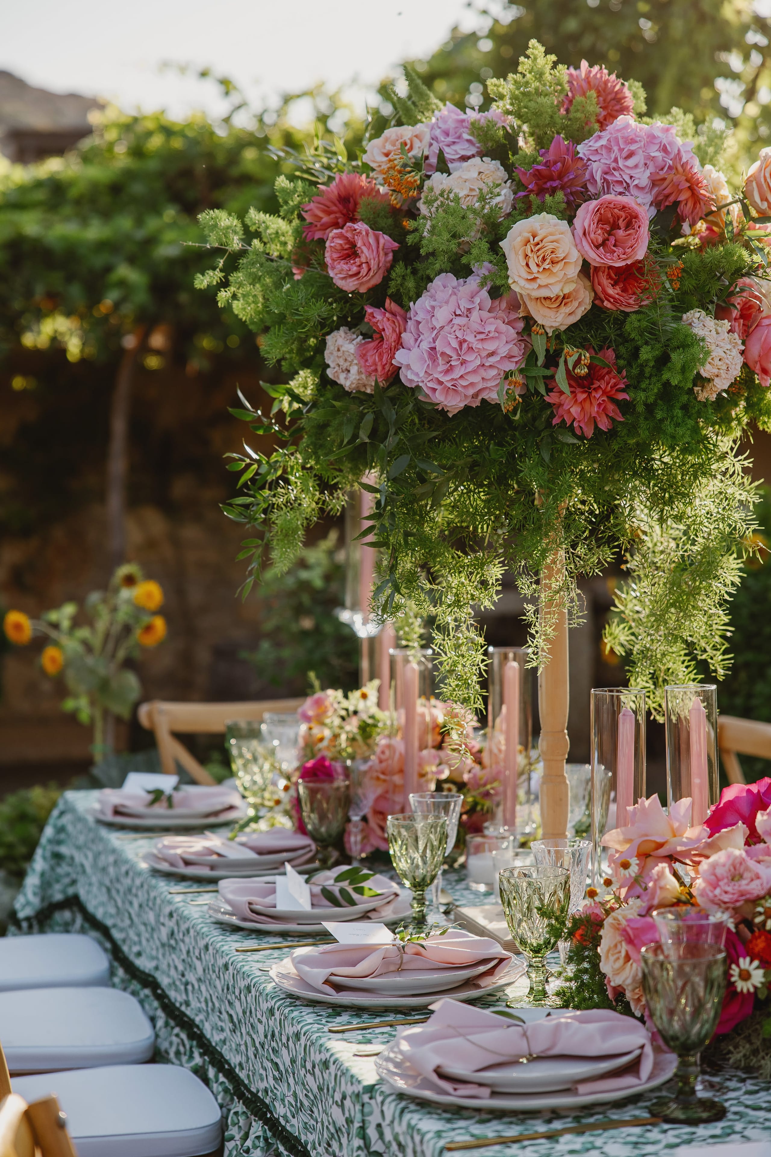 A tall wedding table centrepiece of pink hydrangeas, peach and coral garden roses, and deep pink dahlias, softened by feathery asparagus fern that spills down the stem, set outdoors in Greece. Its pink tones echo the slim pink candles in glass hurricane holders below, and sunflowers glow in the soft-focus background beneath a leafy vine-covered wall.