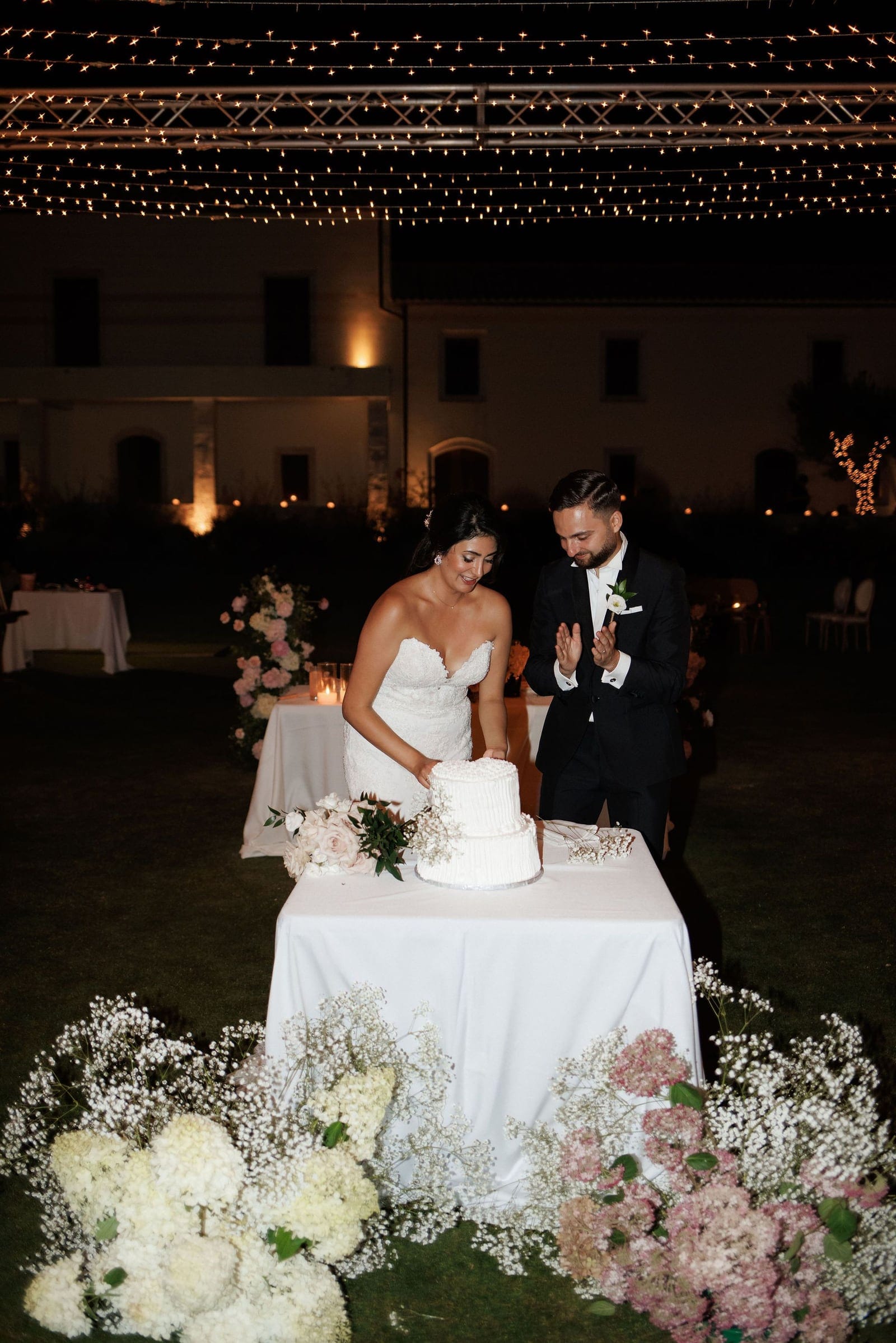 At night on the grounds of Ktima Roxani in Crete, Greece, a bride in a strapless lace gown and a groom in a black suit with a white boutonniere stand at a white-draped table to cut a two-tier white cake. Clusters of white and pink hydrangeas and delicate baby's breath spill across the lawn at the table's base, with a trailing blush-pink floral arrangement beside the cake, all lit by a canopy of warm string lights above.
