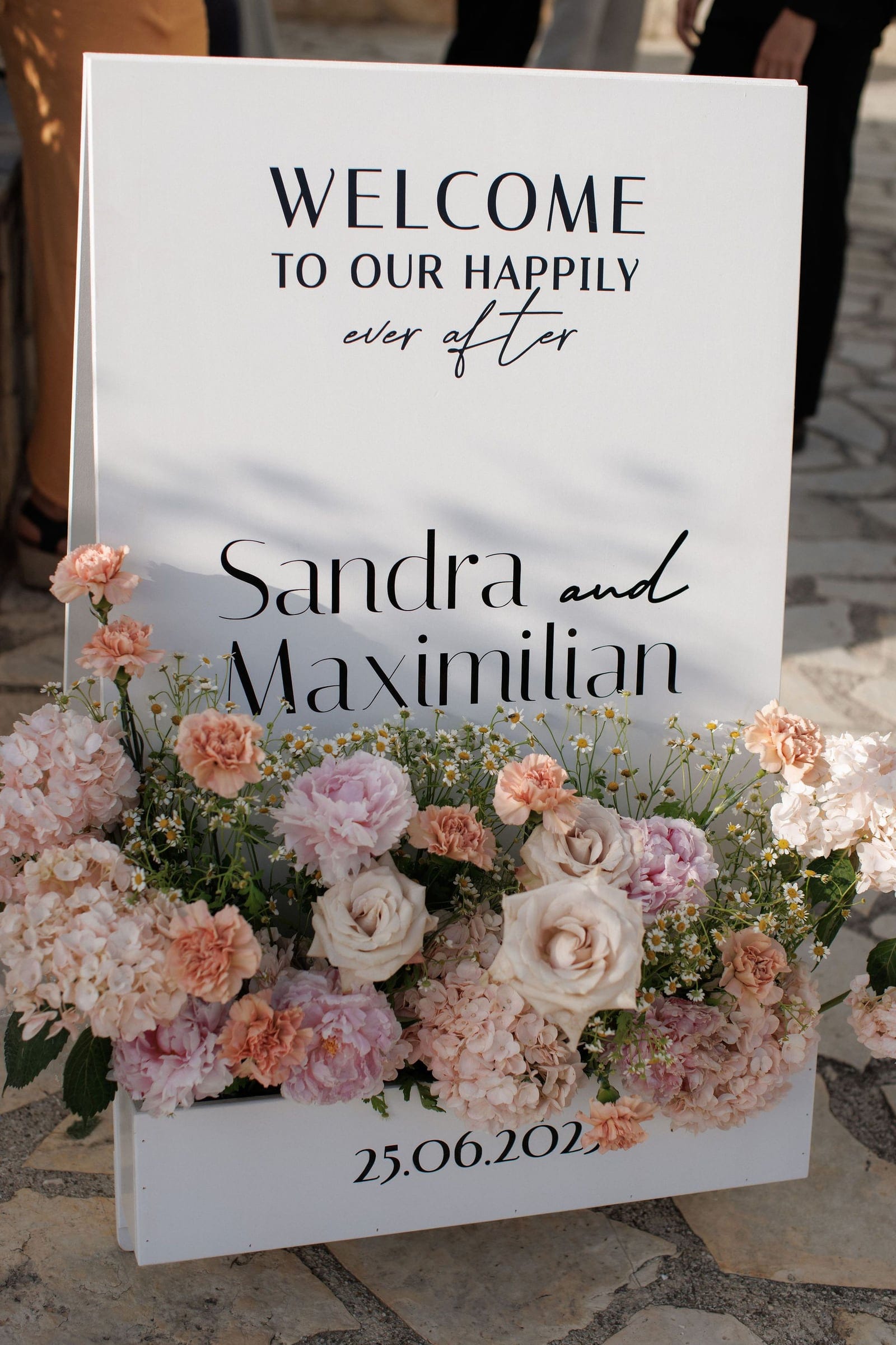A white welcome sign on a stone-paved terrace reads "Welcome to our happily ever after" above the names Sandra and Maximilian, with the date 25.06.202 along the bottom edge. Its base is hidden under a lush wedding arrangement of blush and cream roses, peonies, pale hydrangeas, peach carnations and scattered white chamomile, all in soft pastel tones. This is a wedding floral display at Ktima Roxani, Roxani Estate in Crete, Greece.