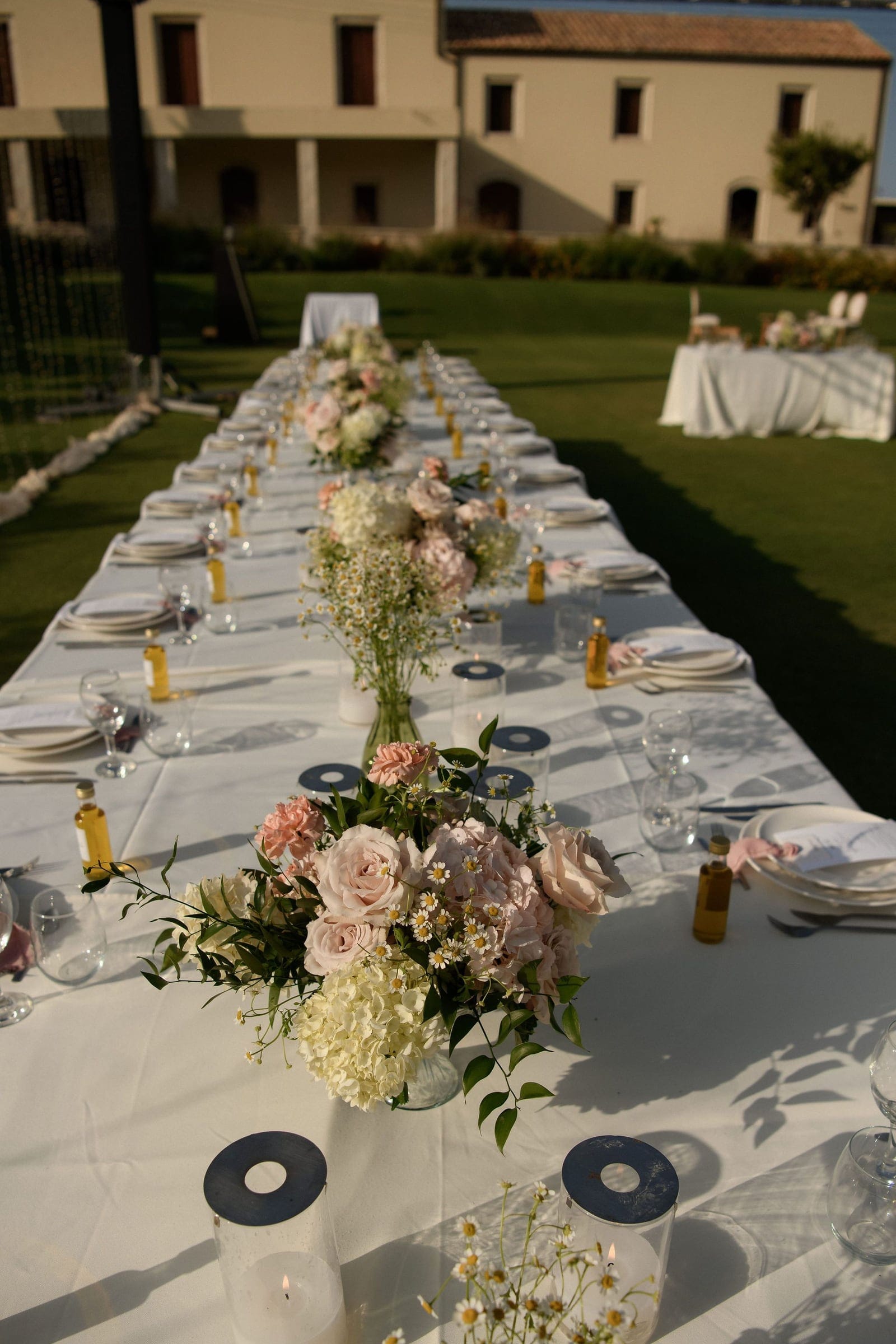 A long outdoor wedding table in Greece, dressed in white linen at Ktima Roxani Estate in Armeni, Crete, with low centrepieces of blush garden roses, pale hydrangeas, carnations and tiny white daisies. Small glass votives with dark round lids sit between them, and amber oil bottles and place settings run down the table, which stretches away across green lawn toward a cream stone building in soft evening light.