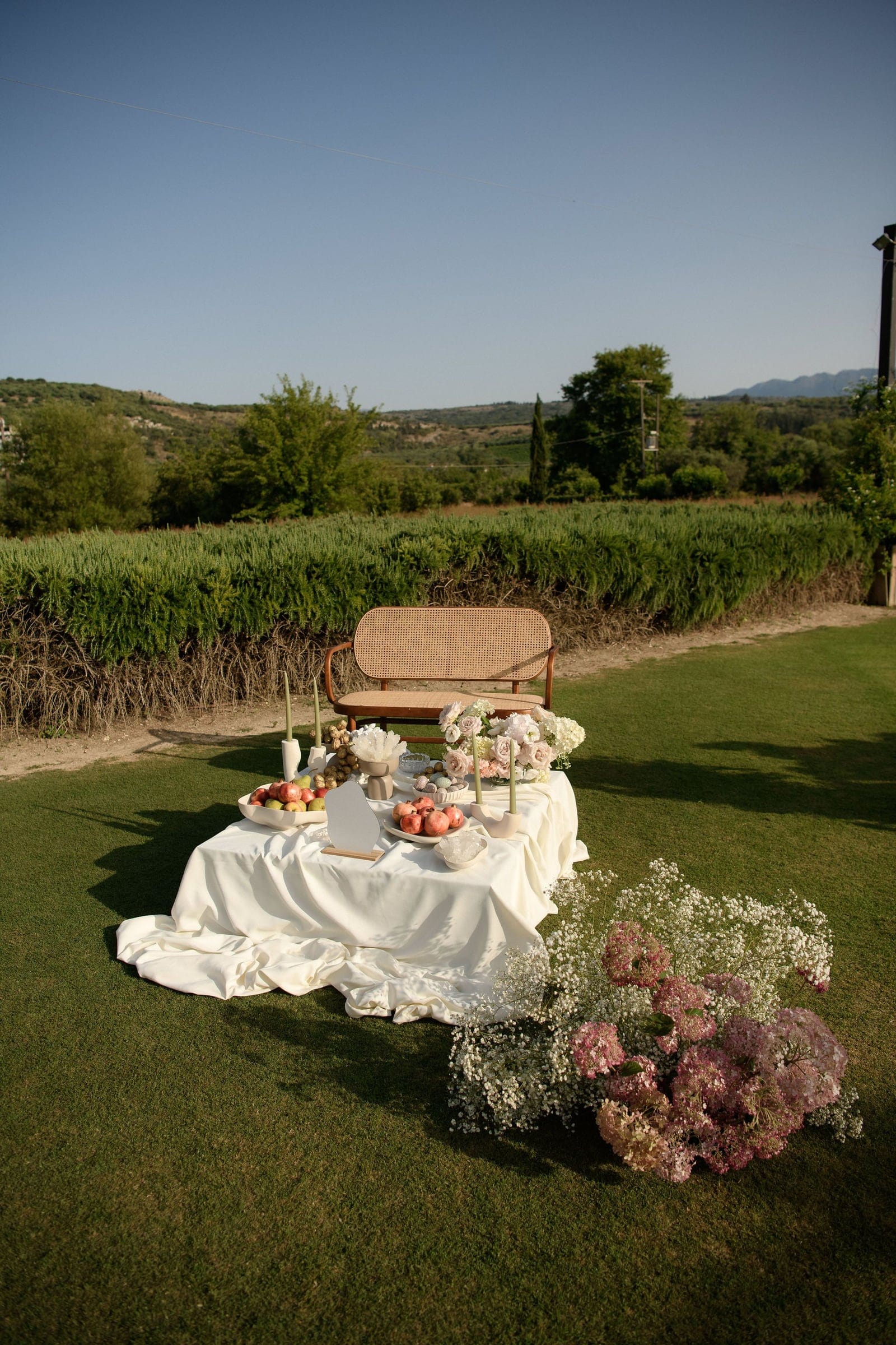A low table draped in cream satin sits on a green lawn at Ktima Roxani Estate in Armeni, Crete, Greece, styled with wedding flowers. Pale pink and white blooms, including roses and hydrangea, sit in a sprawling cluster of gypsophila and dusky pink hydrangea at its front corner, with fruit, pillar candles and a rattan bench set against a hedge and rolling hills under a clear blue sky.