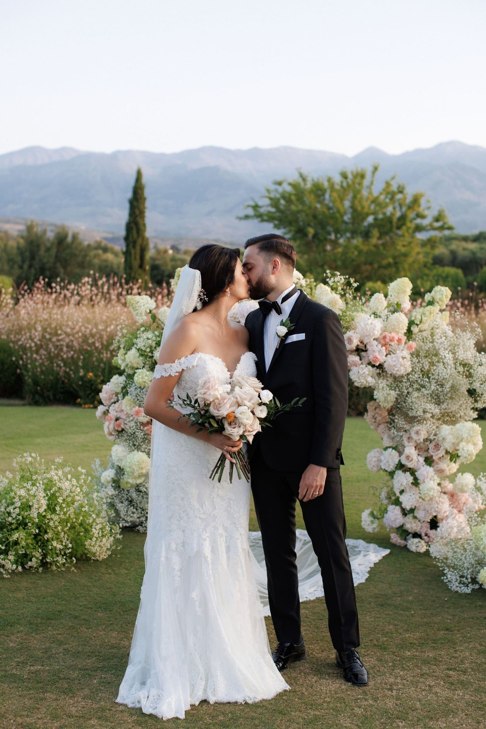 A bride in a lace off-the-shoulder gown with a long veil kisses her groom in a black tuxedo and bow tie, both standing on a lawn in front of a tall wedding arch. The arch is covered in cream and blush roses, white hydrangeas and baby's breath, and she holds a bouquet of soft pink and ivory roses with greenery. Behind them, feathery grasses and olive trees sit under a hazy hillside, lit by warm evening light, all at the Roxani estate in Greece.