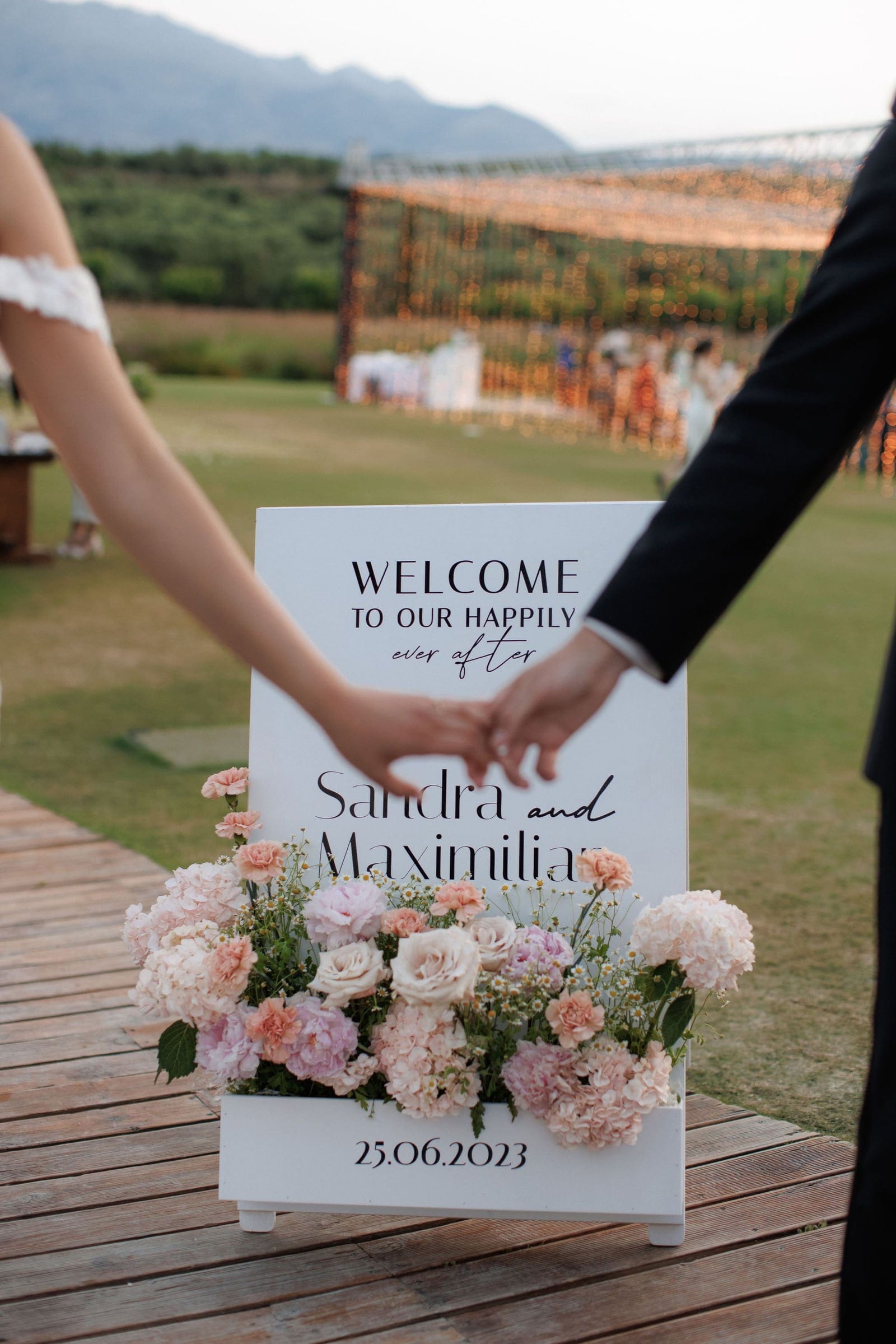 Two hands clasped together beside a white welcome sign reading "Welcome to our happily ever after" with the names Sandra and Maximilian, framed by a planter of blush pink hydrangeas, garden roses, carnations and small white daisies at a wedding at Ktima Roxani in Crete, Greece. Behind them a wooden walkway leads across a green lawn toward a glowing string-light installation at dusk, with mountains softly fading into the sky.
