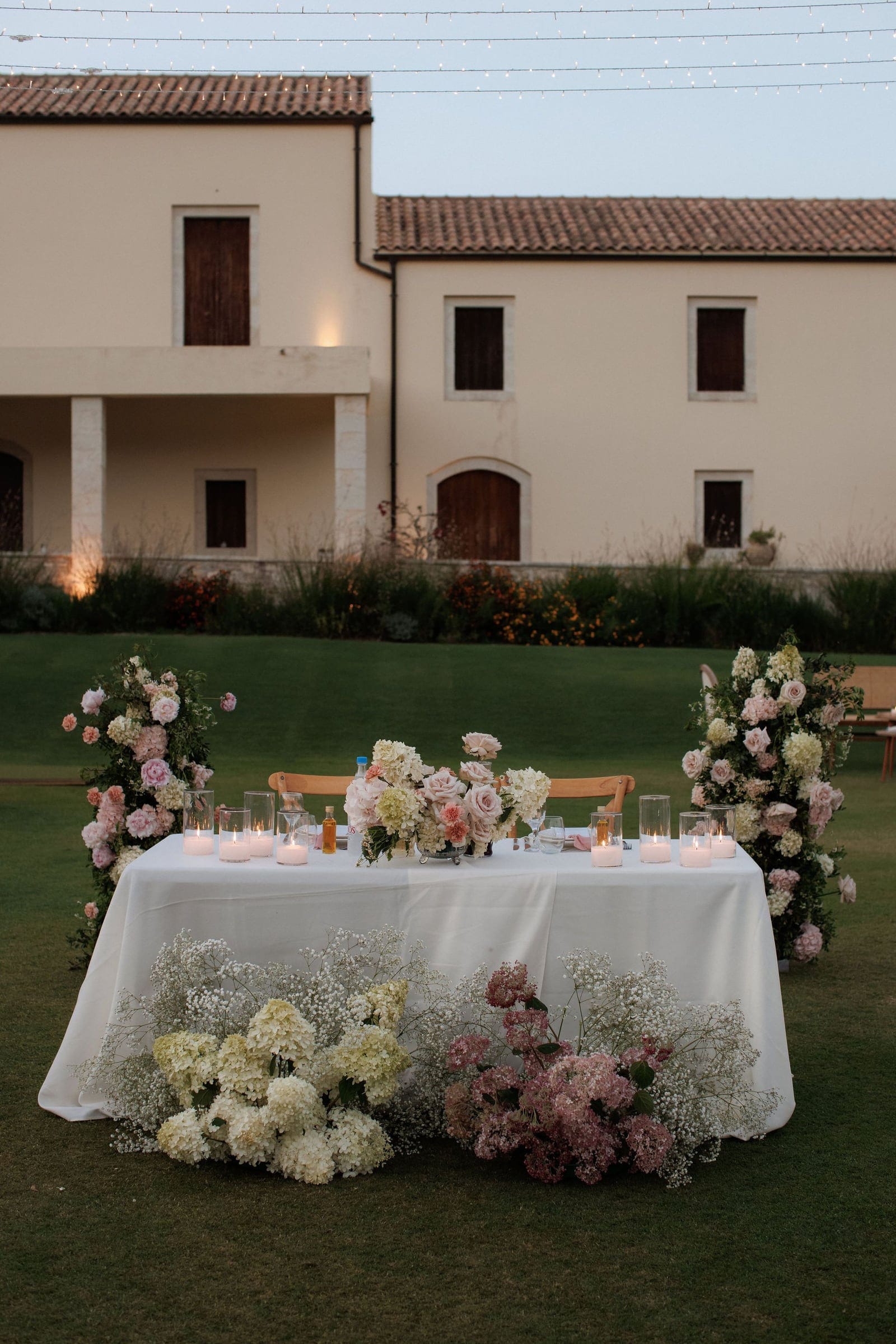 A long white-draped head table stands on a lawn in front of a cream stone villa with terracotta roofs, at dusk. Tall floral arrangements of pink roses, cream and blush blooms rise at each end, with a central low arrangement of roses and white hydrangeas, and pillar candles glow in glass holders along the table. Below the table, drifts of white hydrangeas, dusty pink hydrangeas and delicate baby's breath spill across the grass. This is a wedding table setting at Ktima Roxani Estate in Crete, Greece.