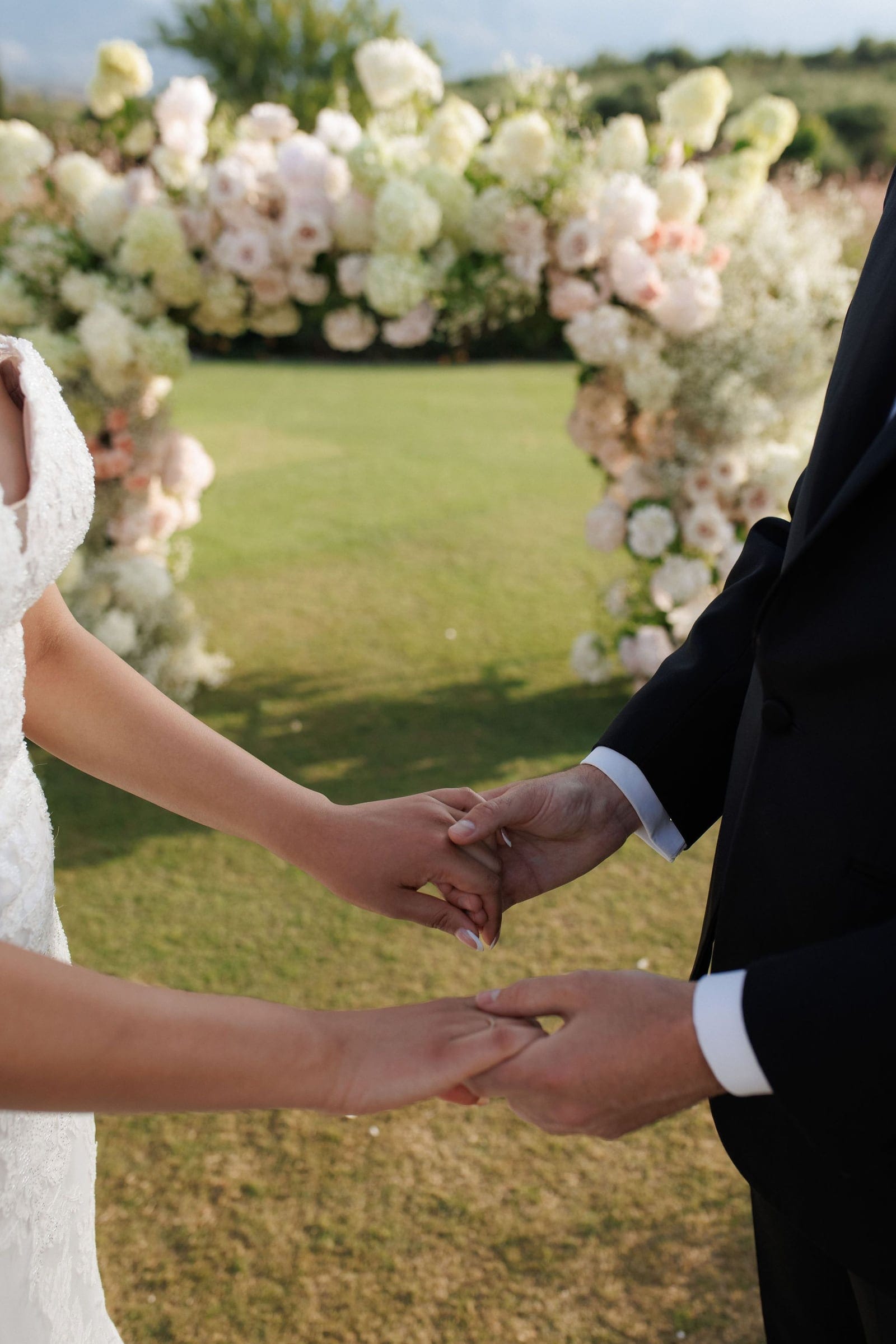 Two pairs of hands clasp together in the foreground, a bride's in lace sleeves and a groom's in a black suit with crisp white cuffs, against a lawn at the Roxani Estate in Armenoi, Crete, Greece. Behind them, a wedding arch is wreathed in cream and blush flowers, including what look like garden roses, hydrangea-like blooms and soft clouds of gypsophila. The soft afternoon light and gentle green lawn make the moment feel tender and intimate.