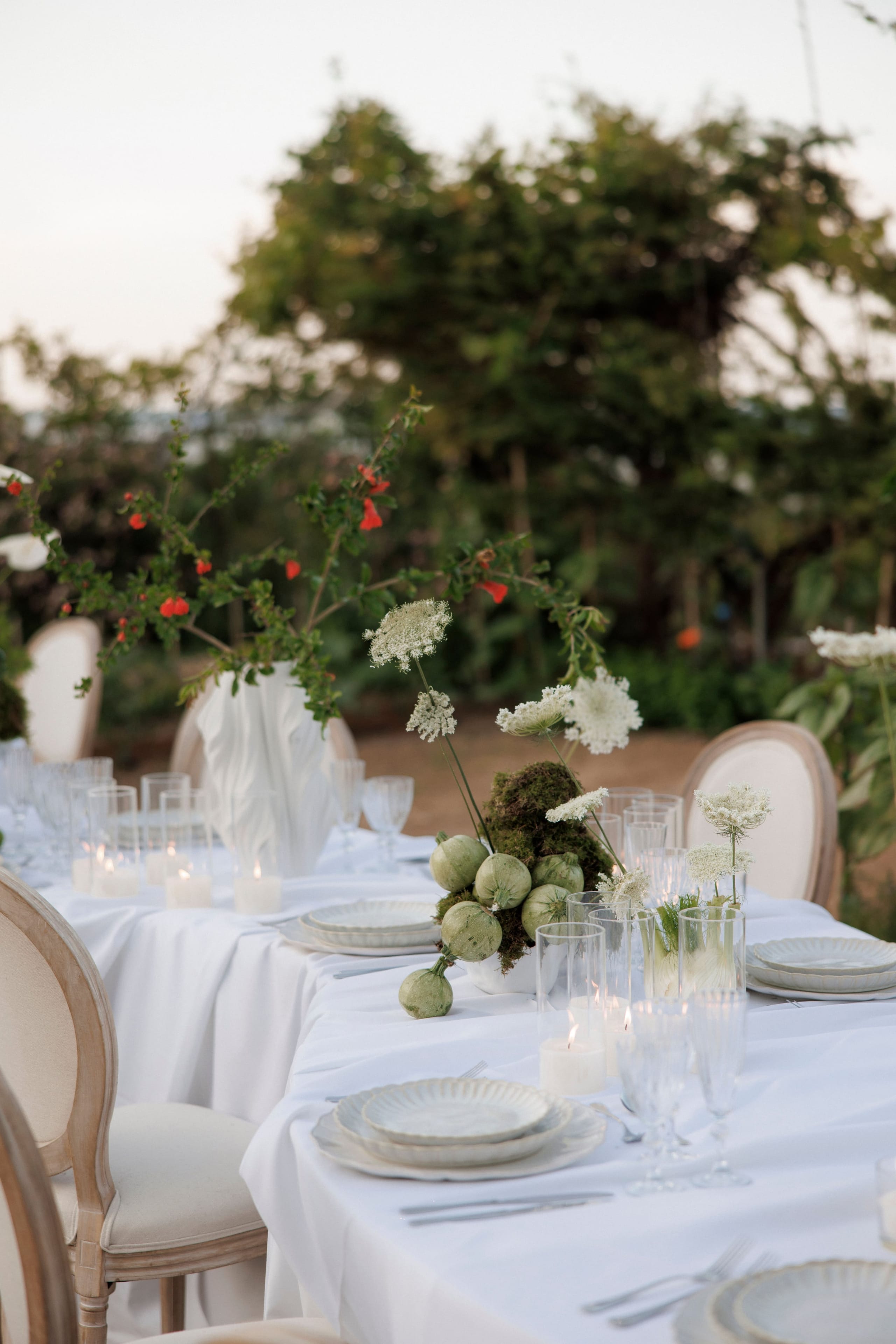 A long table dressed in white linen at dusk, set for a wedding dinner at Gramboussa Restaurant in Kaliviani, Crete, Greece. In the foreground, a low arrangement of green seed pods and moss sits beside sprays of white Queen Anne's lace, while a tall white vase holds trailing branches with small red flowers. Cream-backed chairs line the table, with stemmed glasses, lit candles in glass and pale plates, and soft green foliage blurred behind.