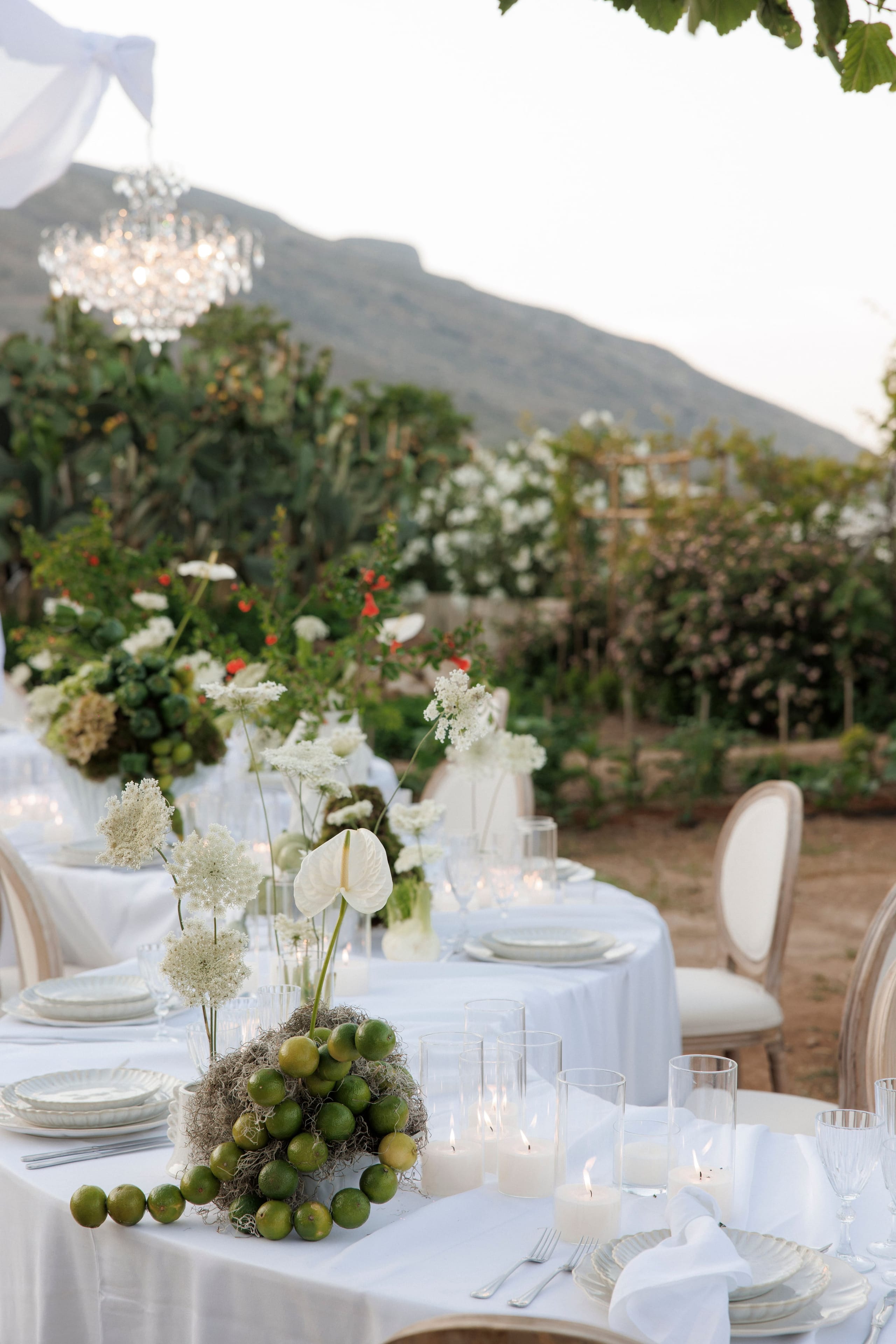 A long round dining table dressed in white linen on an open-air terrace at Gramboussa Restaurant in Kaliviani, Crete, Greece, set for a wedding reception. The centrepiece is a low nest of green limes wrapped in moss, with a white anthurium and sprays of white Queen Anne's lace rising beside it, while white candles glow in glass hurricanes and a crystal chandelier hangs above under the evening sky.